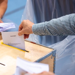 Två personer lägger valsedlar i en valurna av trä. En röstar med ett valkuvert och en håller en bunt kuvert.