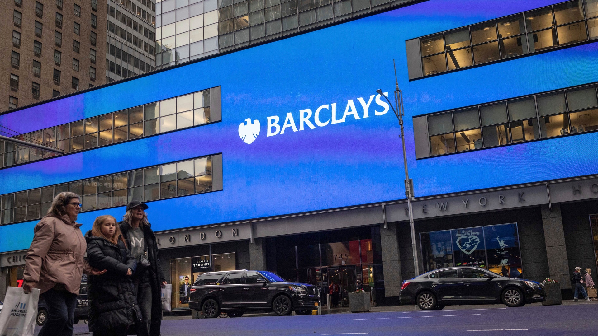 En stadsgata framför en modern byggnad med en stor blå Barclays-skylt. Några personer går på trottoaren framför byggnaden.