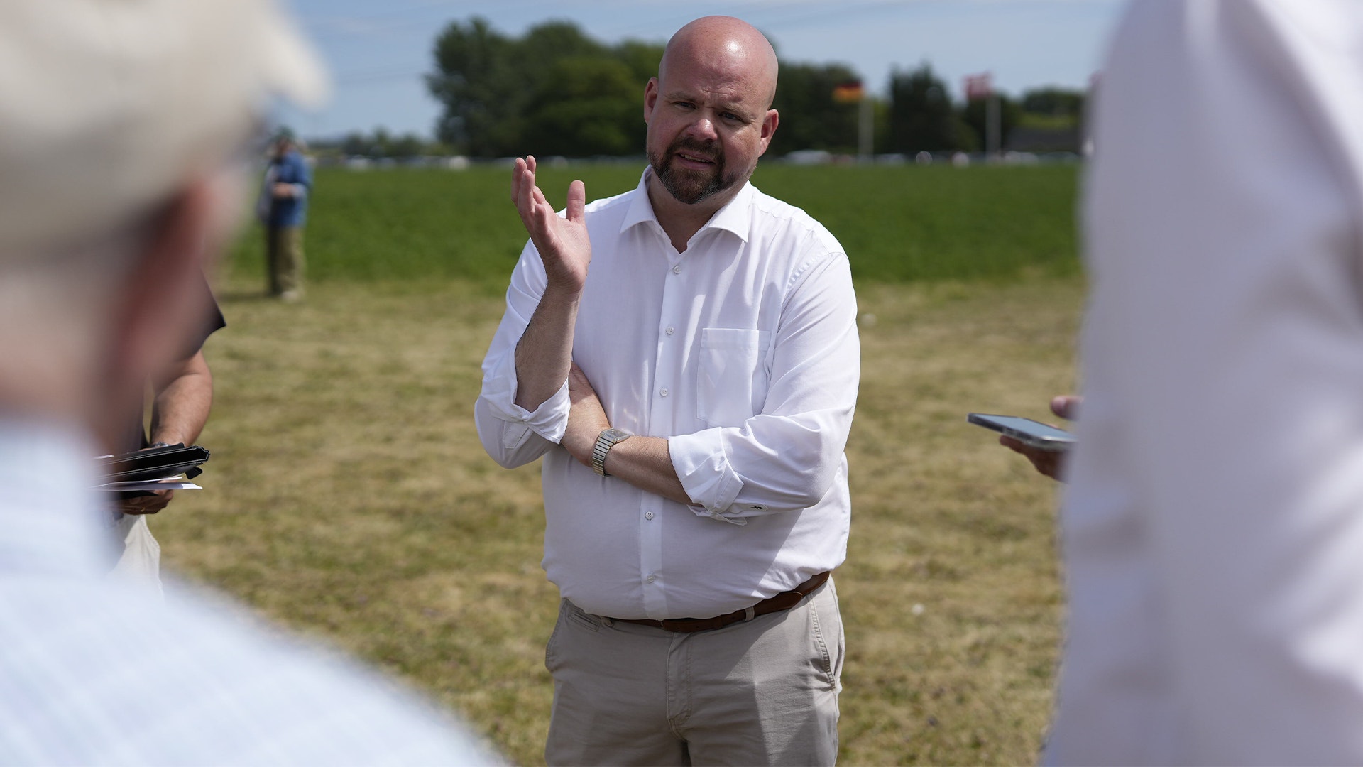 En man i vit skjorta talar och gestikulerar på en gräsyta med landskap i bakgrunden.