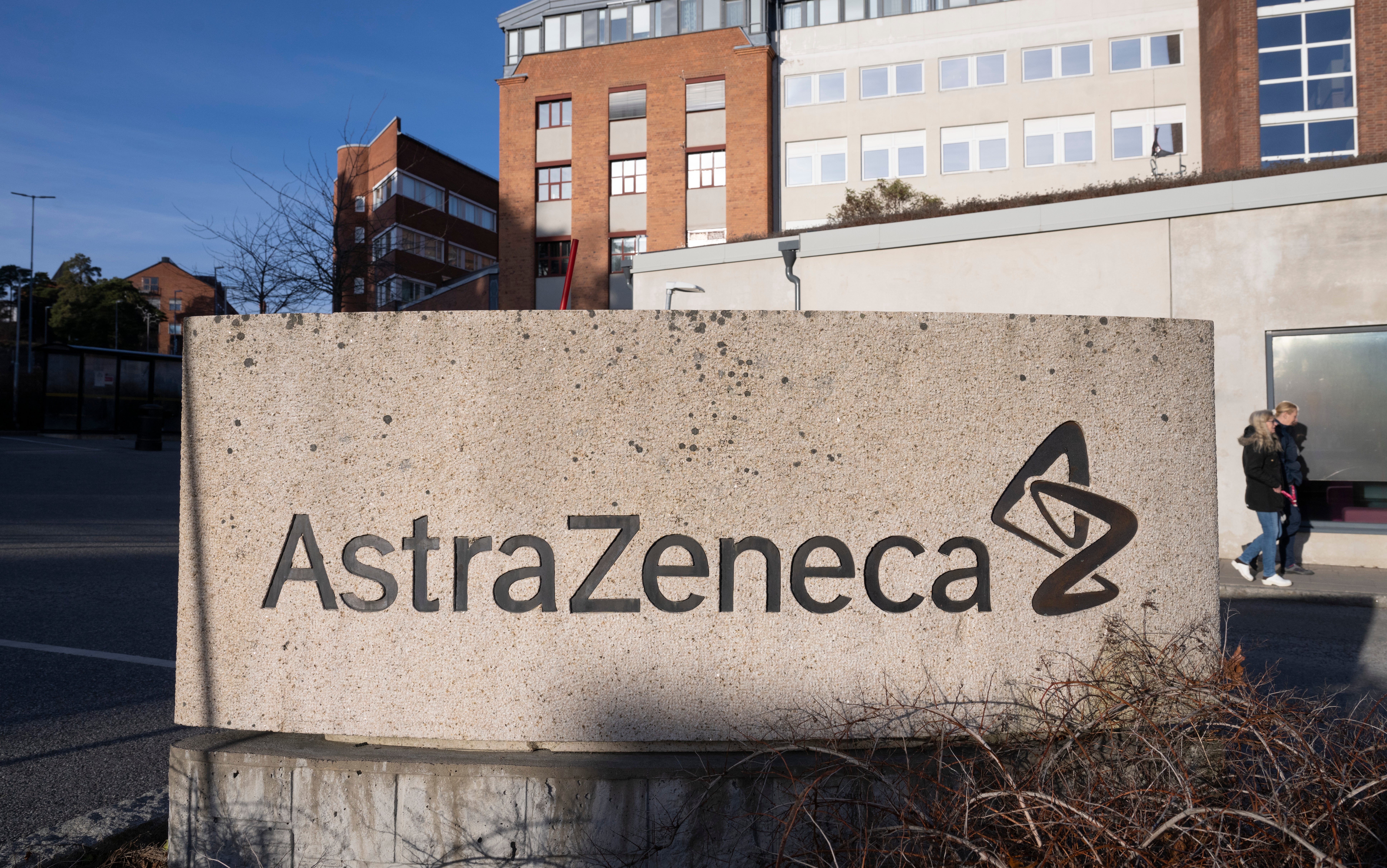 Betongvägg med AstraZeneca-logotypen vid företagets anläggning, med kontorsbyggnader i bakgrunden.
