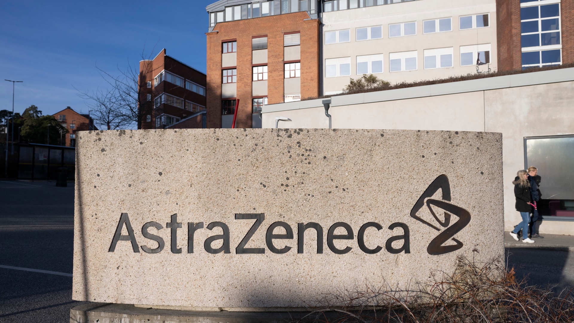 Betongvägg med AstraZeneca-logotypen vid företagets anläggning, med kontorsbyggnader i bakgrunden.