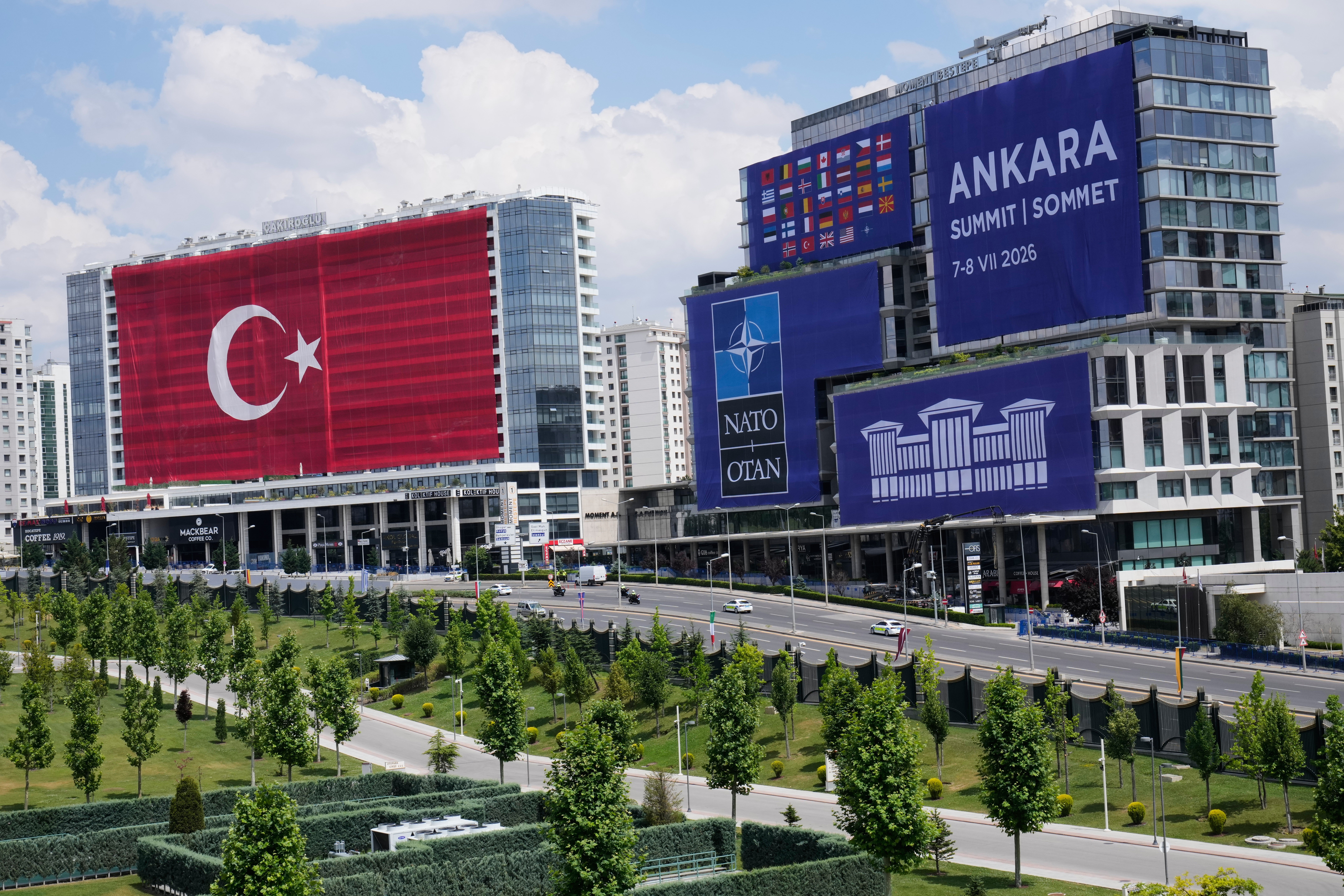 Stadsvy från Ankara med moderna byggnader dekorerade med turkisk flagga och NATO-symboler. En grön park med träd ligger i förgrunden.