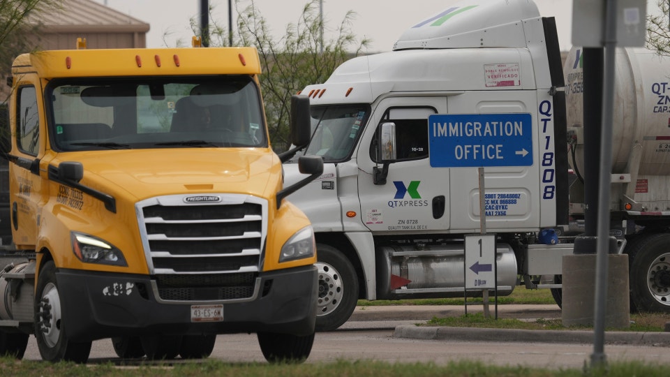 En gul lastbil står parkerad bredvid en vit lastbil. Bilarna är placerade på en parkeringsplats med träd och gräs i bakgrunden.