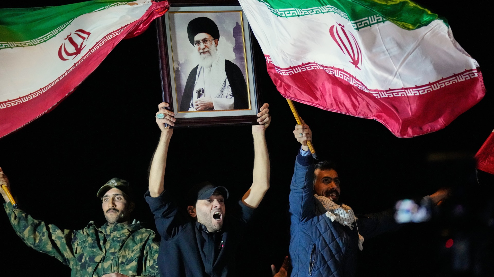 Demonstranter håller upp iranska flaggor och en porträttbild under en nattlig samling.