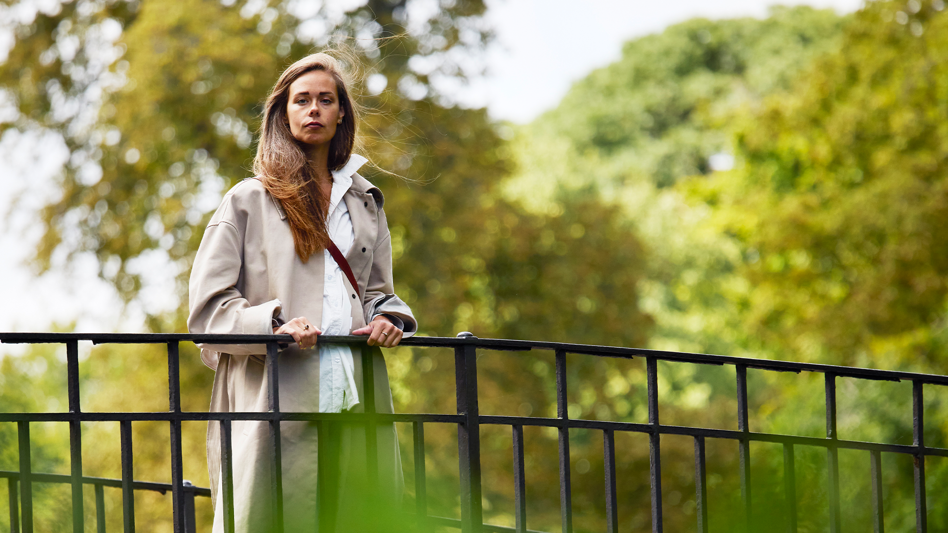 Julia Ravanis står i grönsken på en bro över en älv. Hon har på sig en beige trenchcoat och det blåser i håret.
