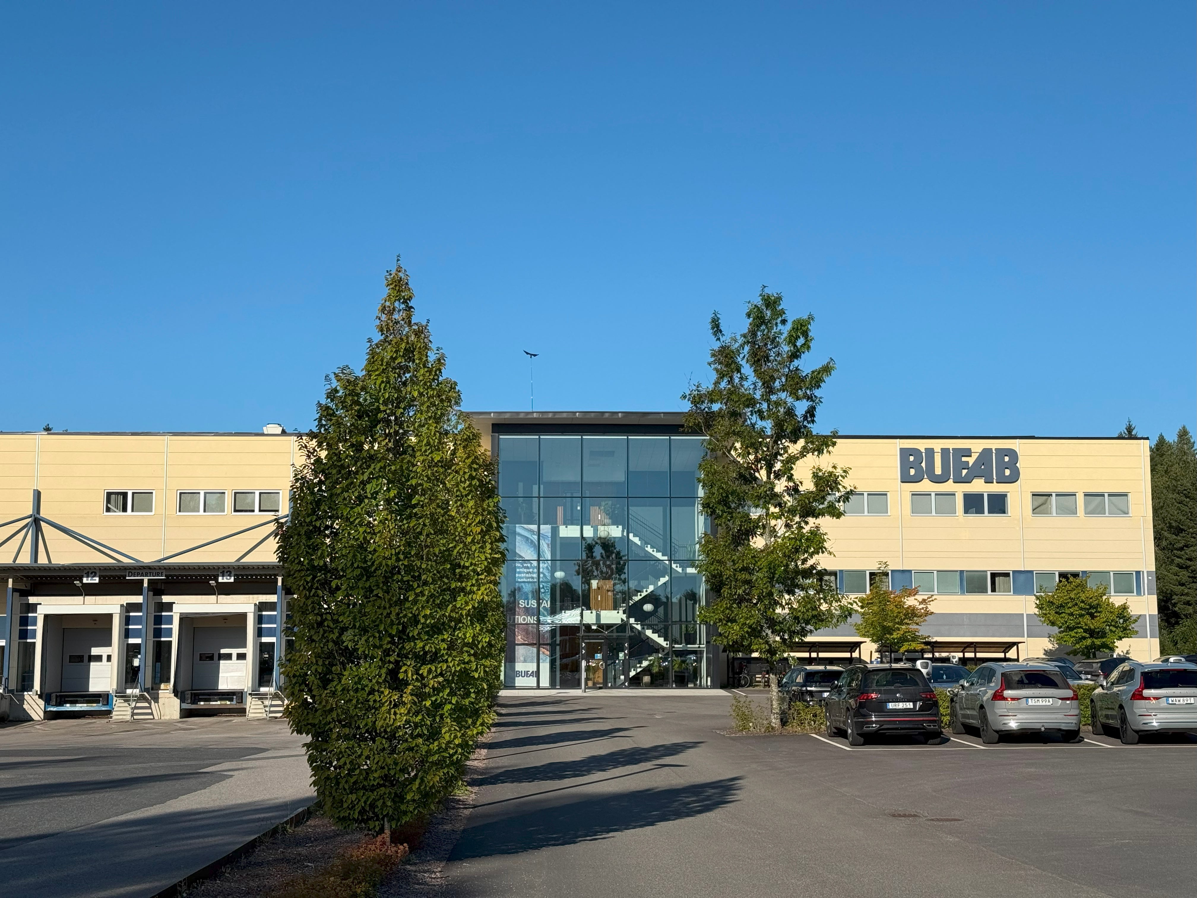 Modernt gult kontorsbyggnad med BUF4B-skylt, parkerade bilar och gröna växter framför entrén på en solig dag.