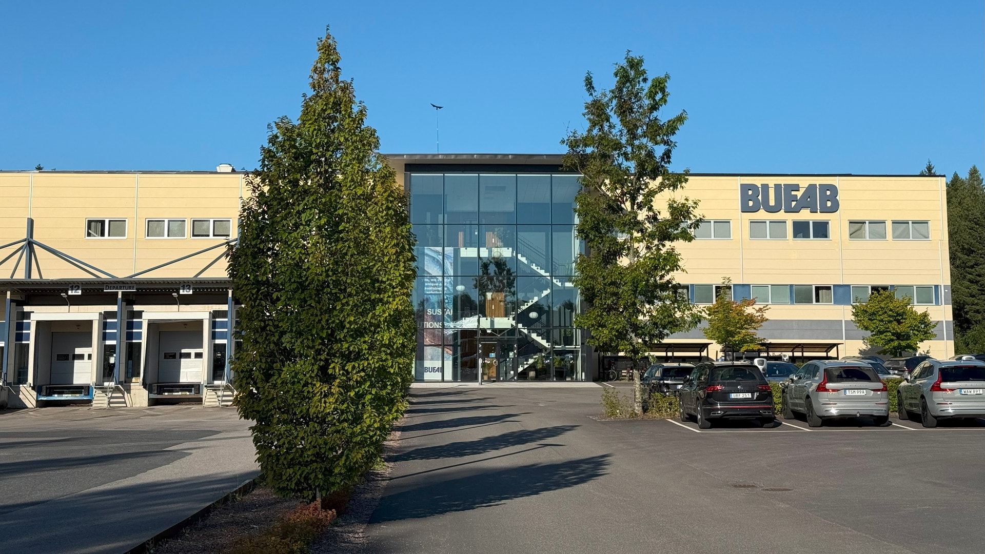 Modernt gult kontorsbyggnad med BUF4B-skylt, parkerade bilar och gröna växter framför entrén på en solig dag.