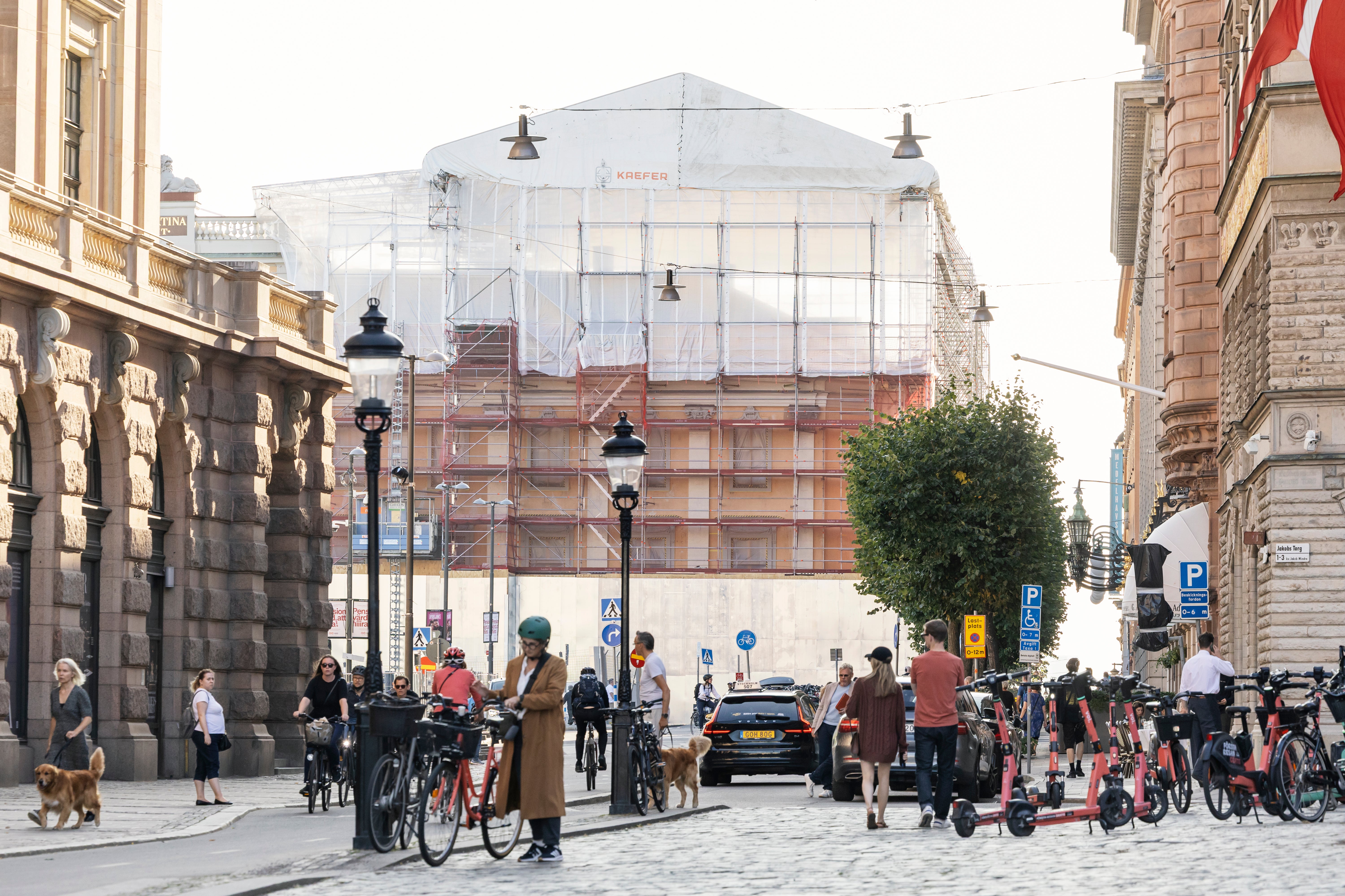 En stadsgata med klassiska byggnader, fotgängare, cyklister och en byggnad under renovering med vita ställningar.