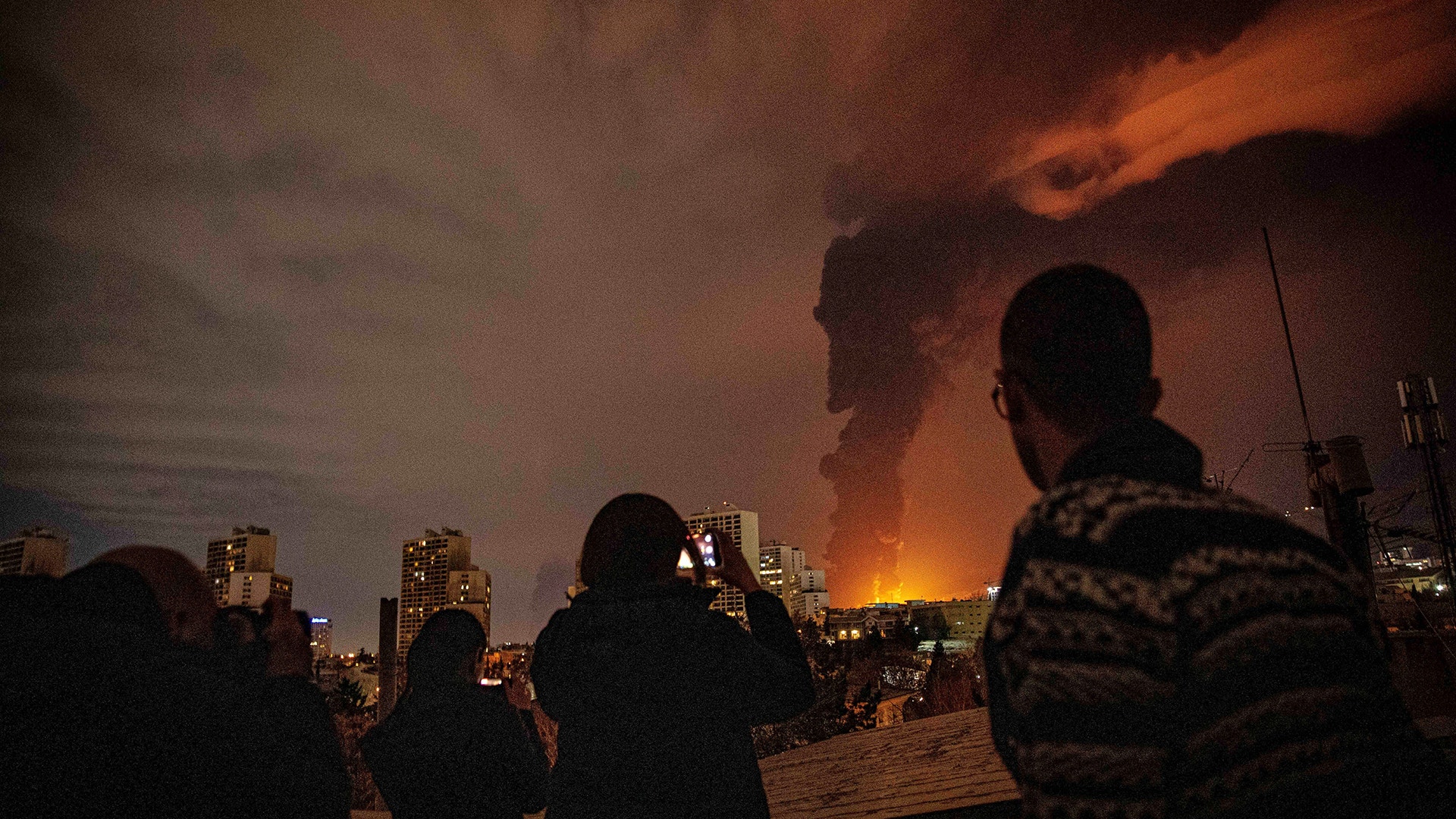 Silhuetter av människor som observerar en stor brand eller explosion i en stad på natten, med orange lågor och rök som lyser upp himlen.