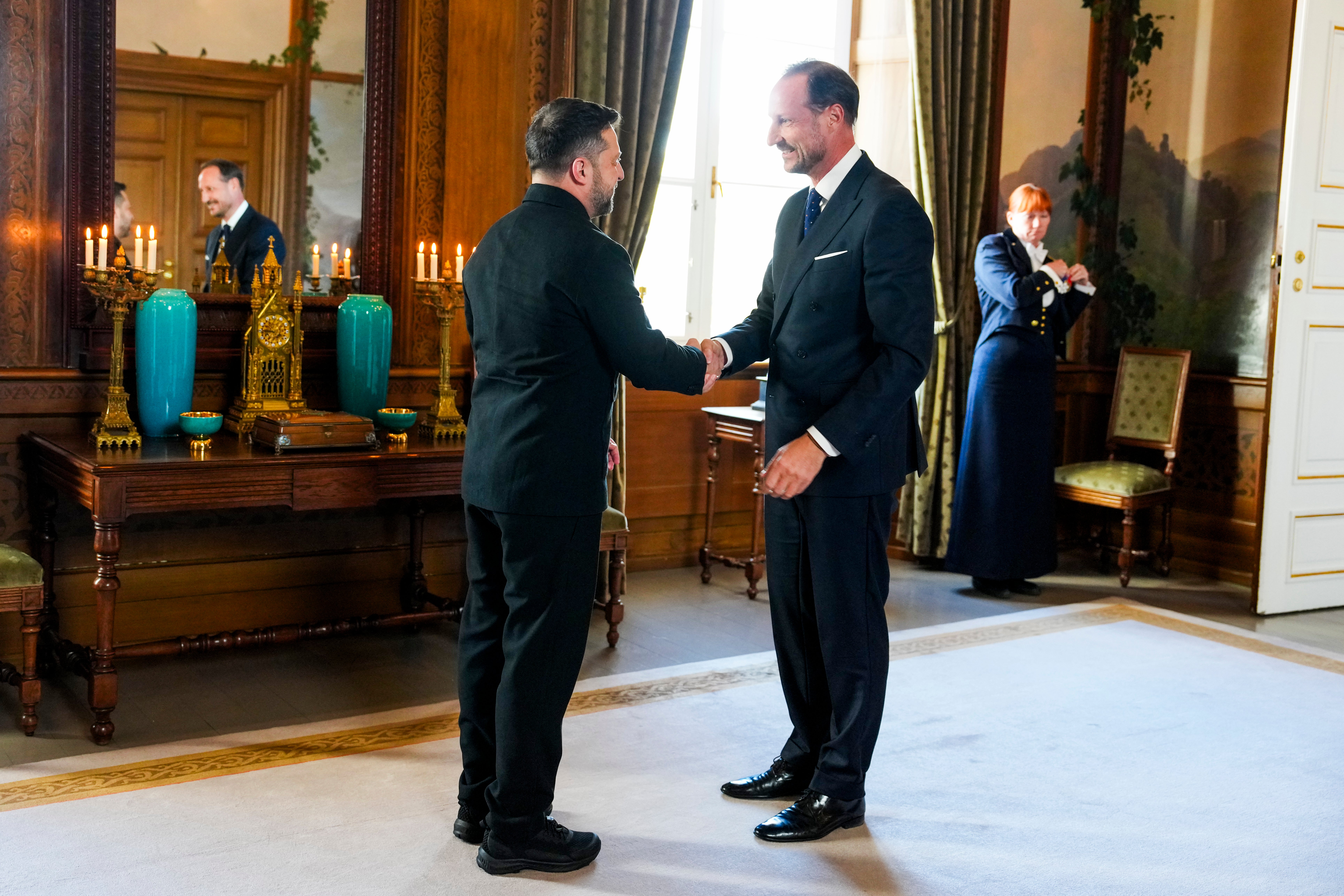 Två män i mörka kläder skakar hand i ett festligt inrett rum på ett slott.