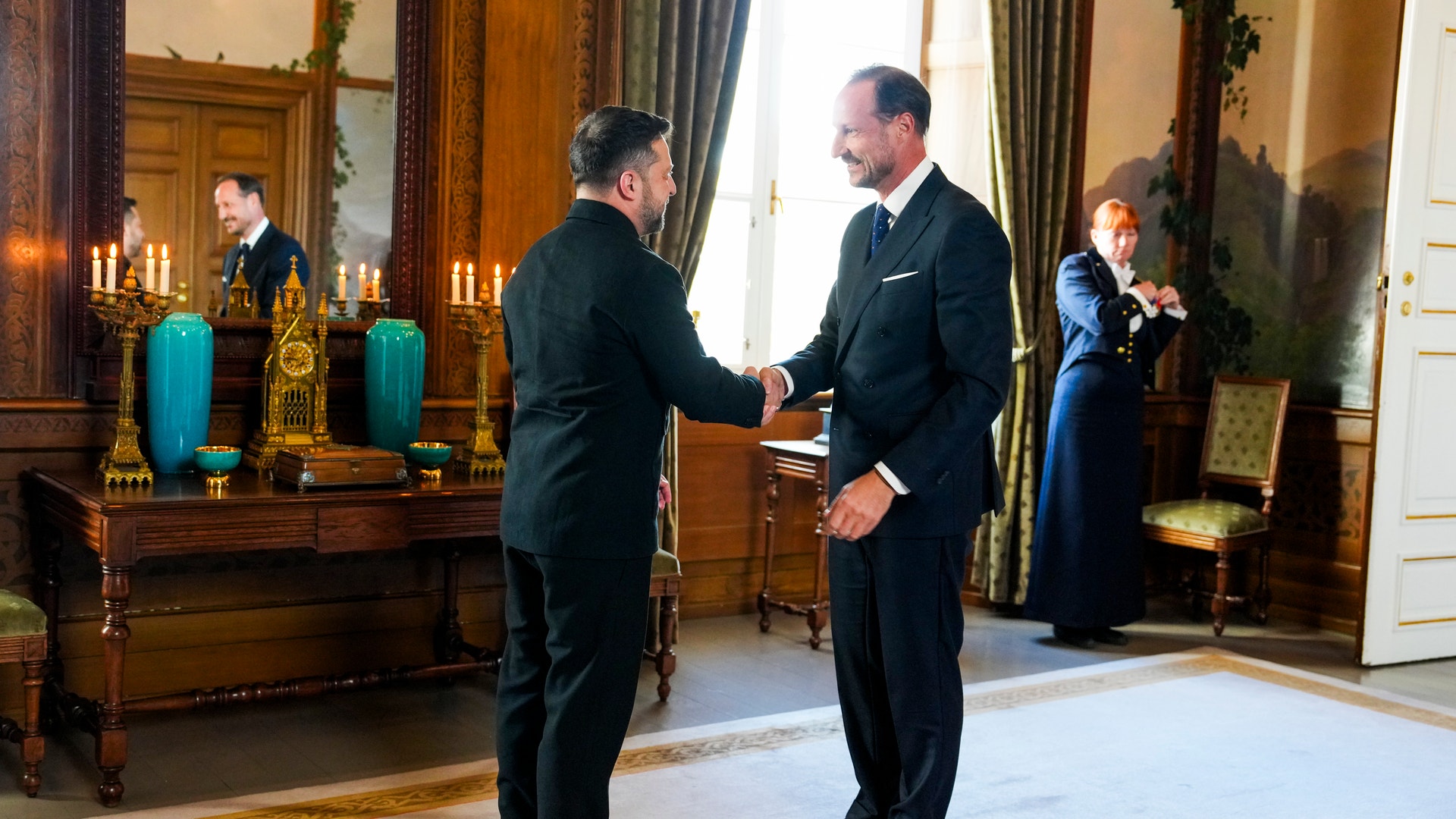 Två män i mörka kläder skakar hand i ett festligt inrett rum på ett slott.