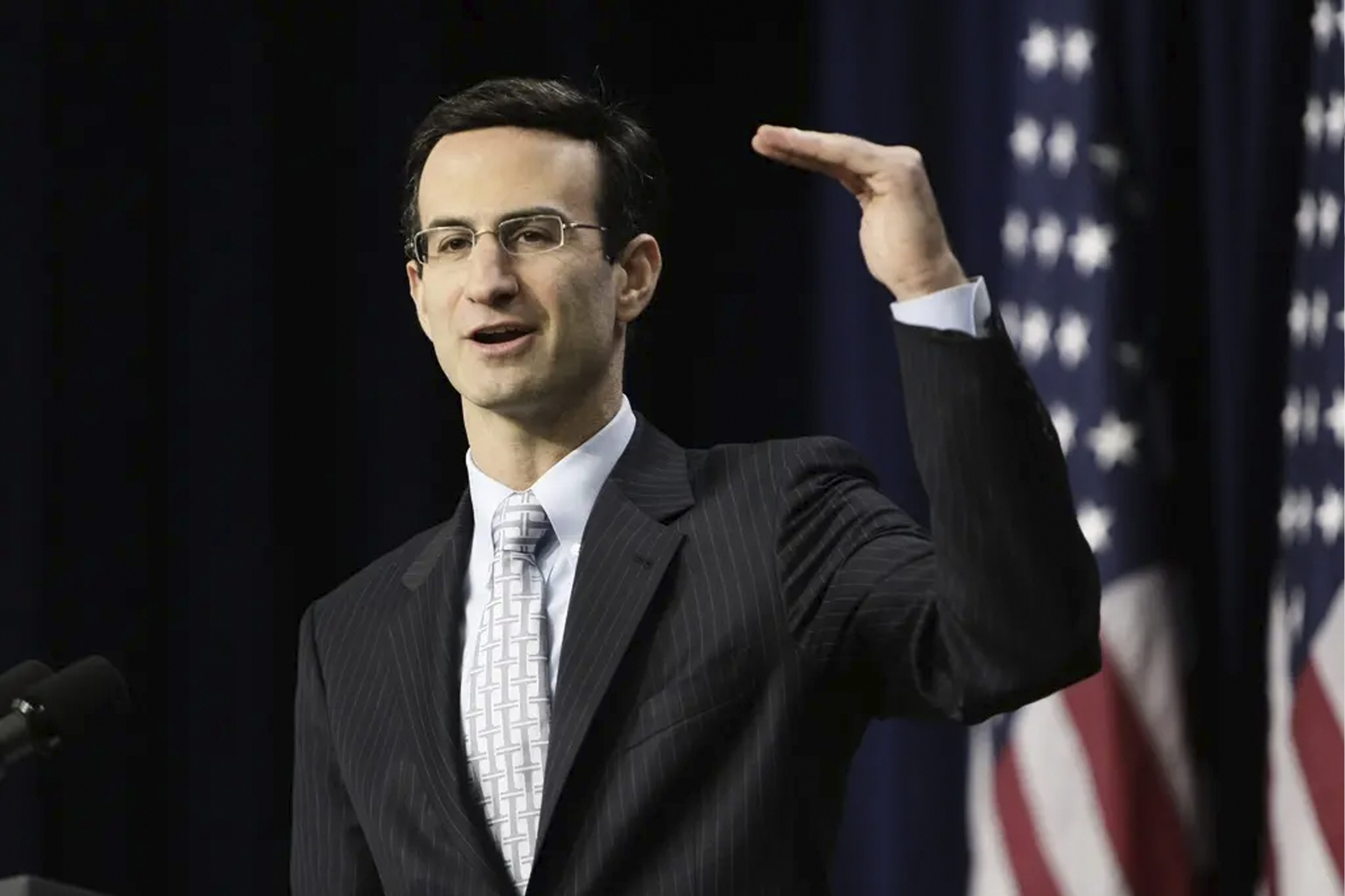 Man i kostym och glasögon håller tal framför amerikansk flagga.