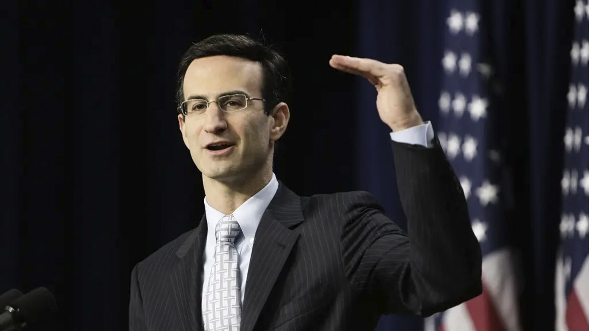 Man i kostym och glasögon håller tal framför amerikansk flagga.