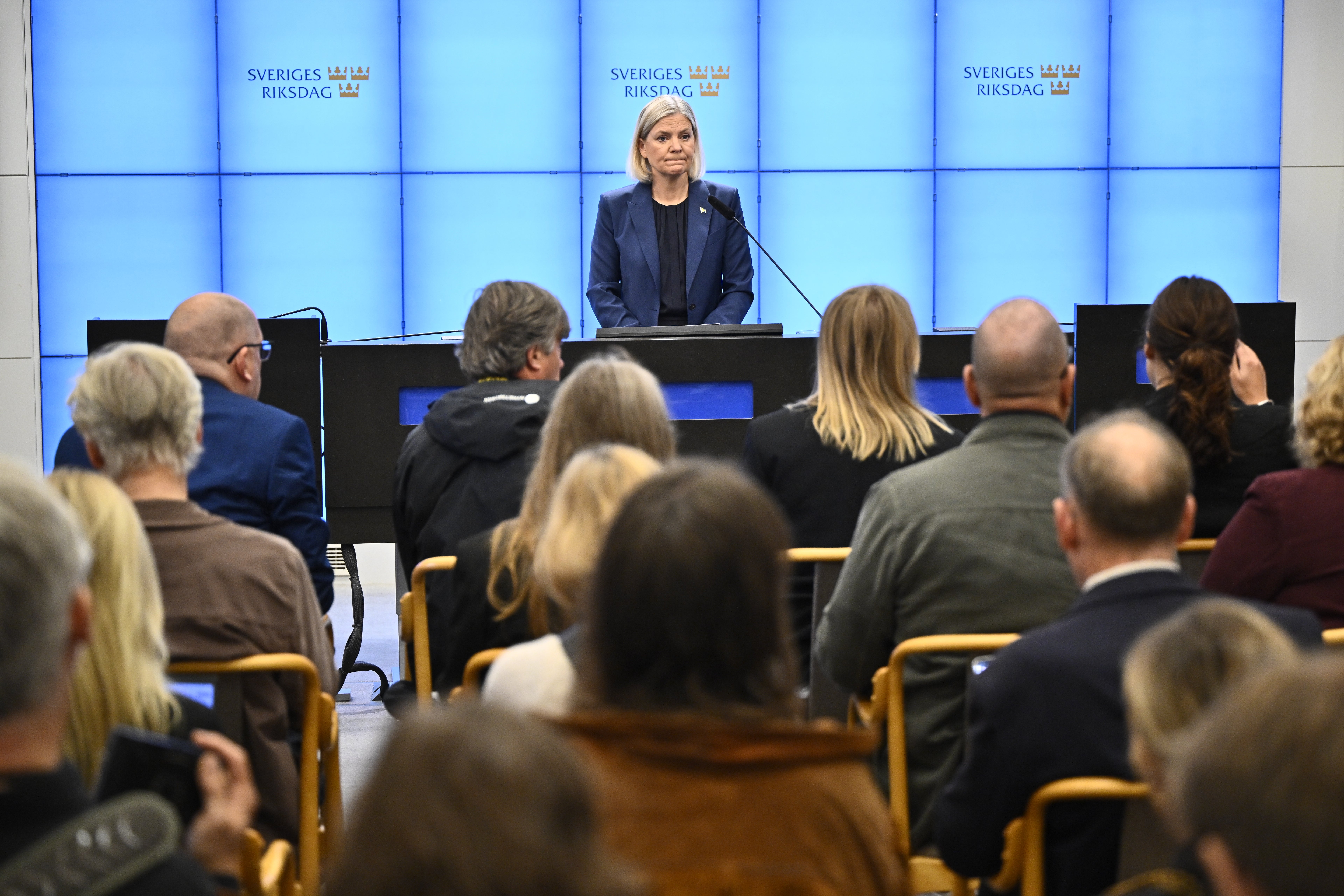En kvinna talar vid ett podium framför publik, med skylten Sveriges Riksdag i bakgrunden.