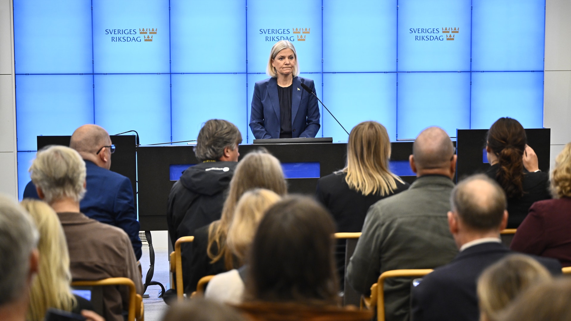 En kvinna talar vid ett podium framför publik, med skylten Sveriges Riksdag i bakgrunden.