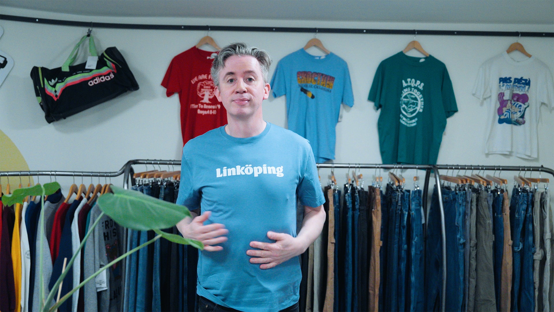 Fredrik Strage i en ljusblå t-shirt som det står Linköping på, med massa kläder som hänger på galgar i bakgrunden.