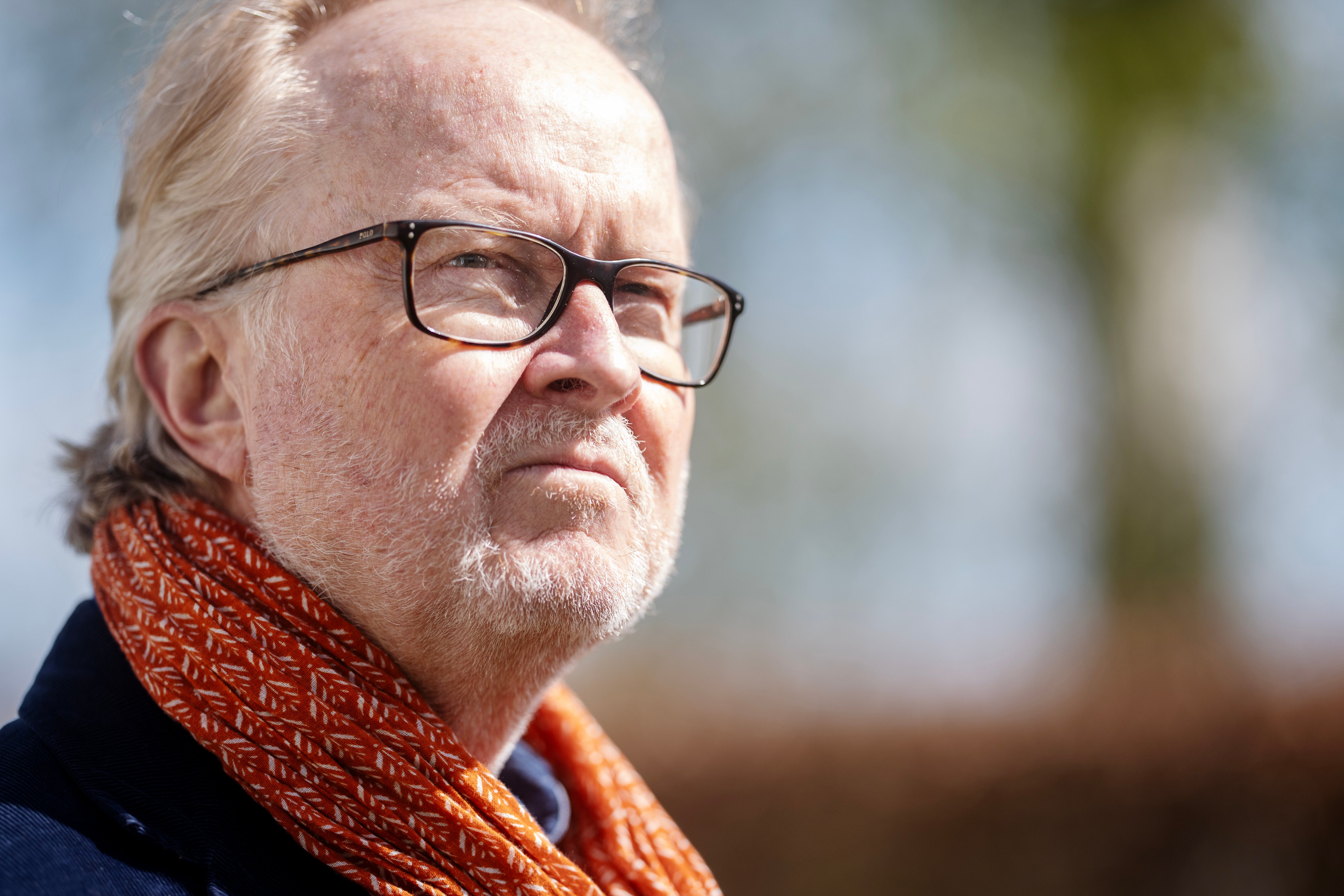 Porträtt av en äldre man med vitt hår och glasögon, iklädd mörkblå jacka och röd-orange halsduk.