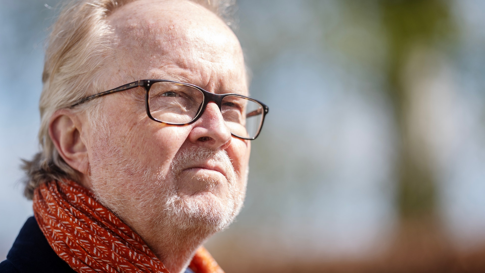 Porträtt av en äldre man med vitt hår och glasögon, iklädd mörkblå jacka och röd-orange halsduk.
