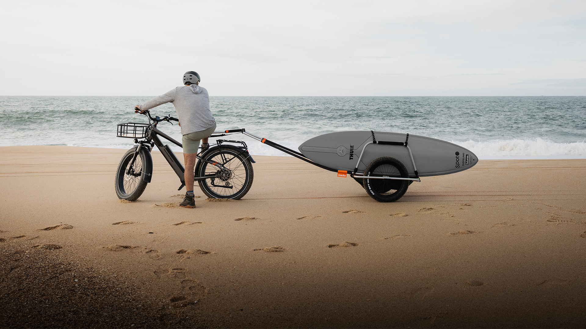 En person cyklar med en elcykel på en strand och drar en surfbräda på en cykelkärra.