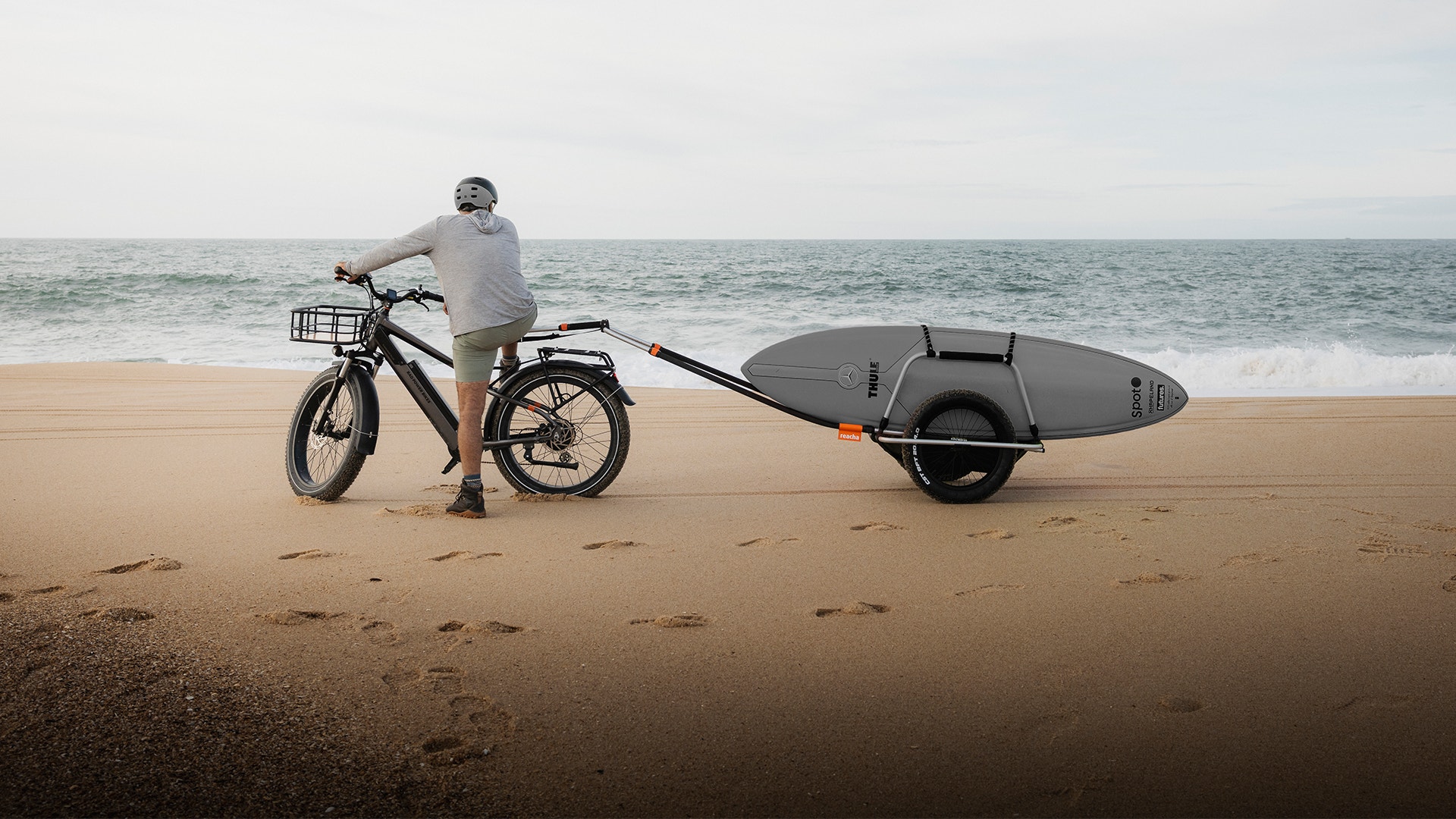 En person cyklar med en elcykel på en strand och drar en surfbräda på en cykelkärra.
