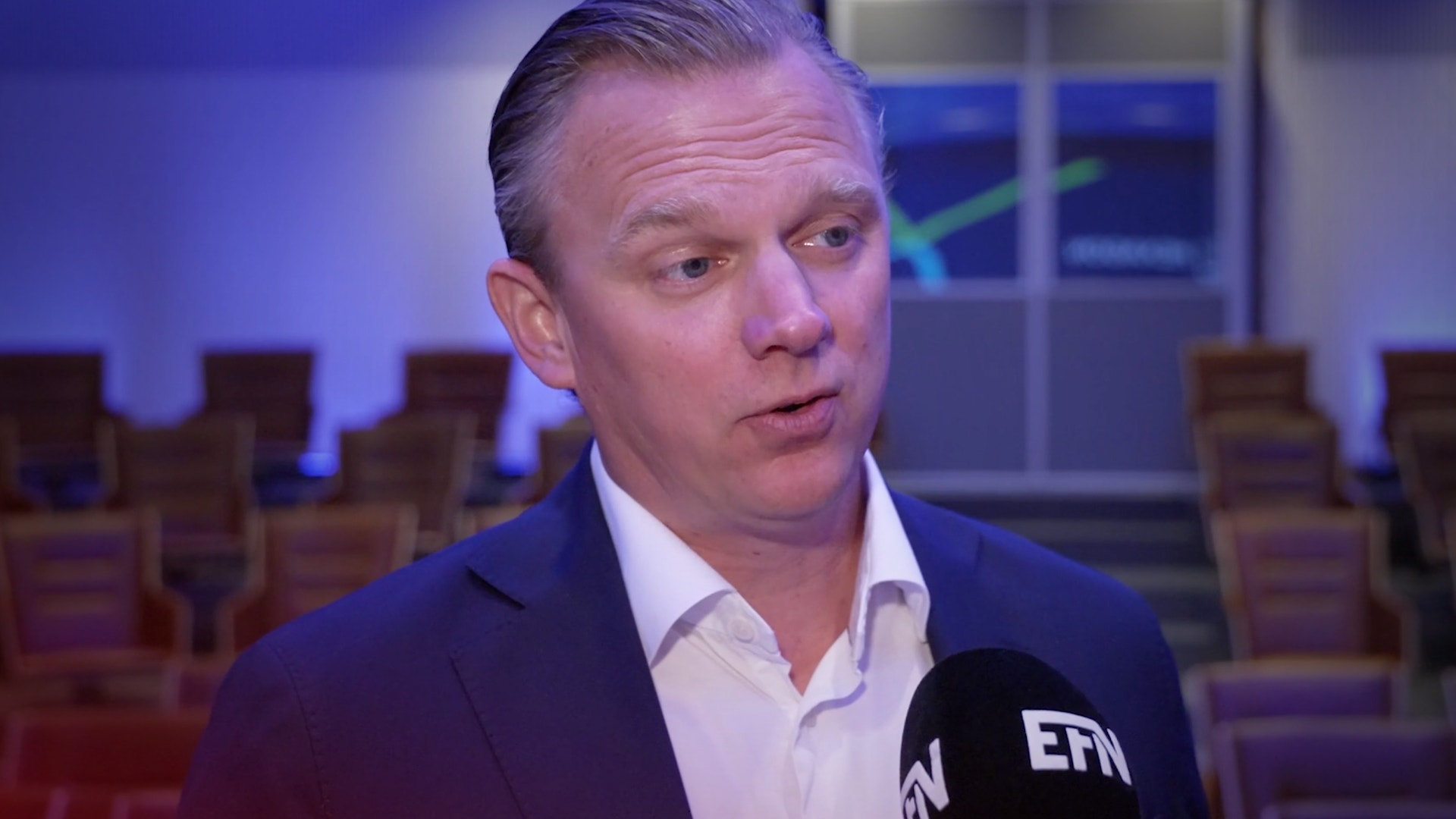 Man i mörkblå kostym blir intervjuad med mikrofon i en konferenssal med blå belysning.
