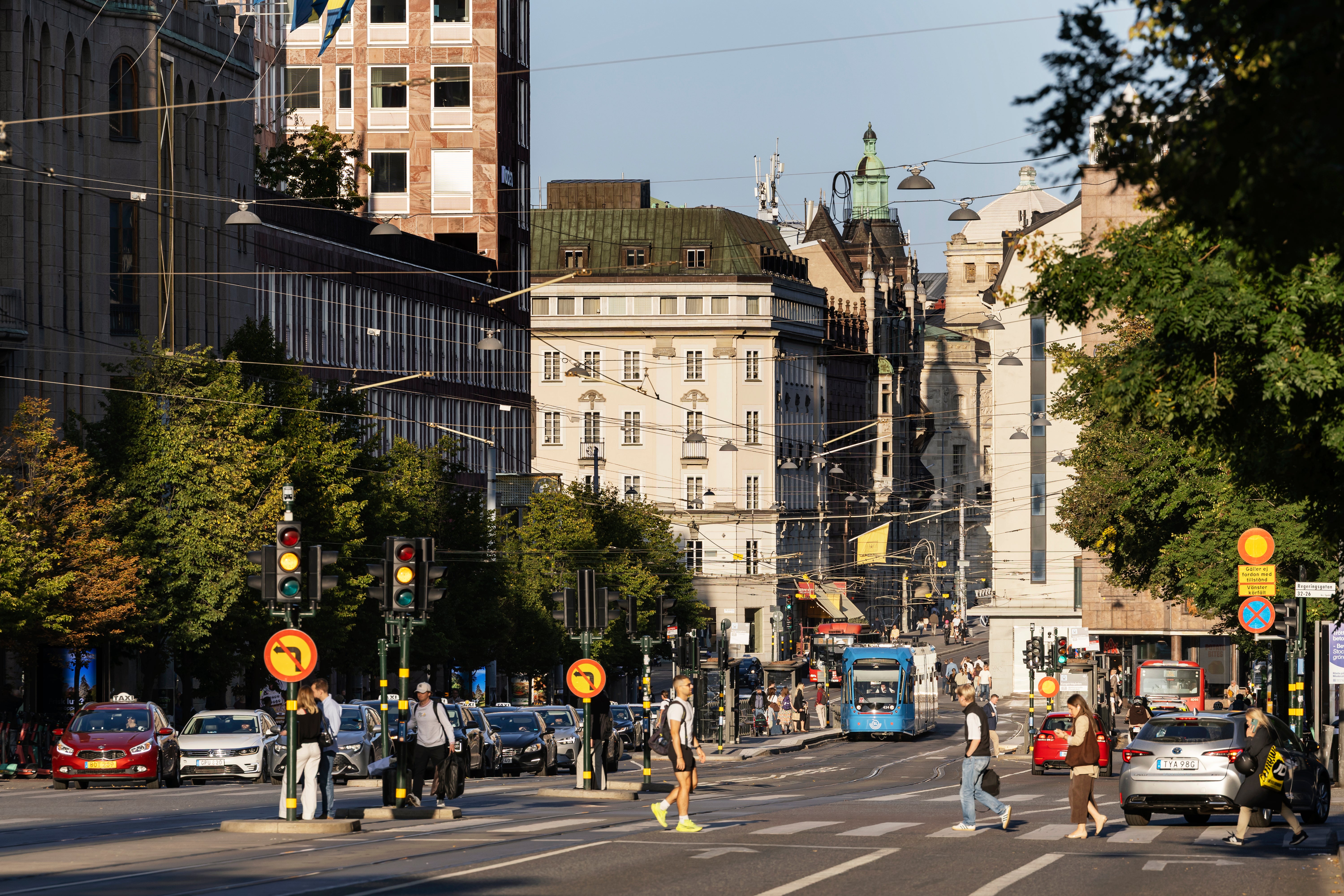En stadsgata i en europeisk stad med klassiska byggnader, gågränd med fotgängare, trafikljus och gröna träd längs vägen.