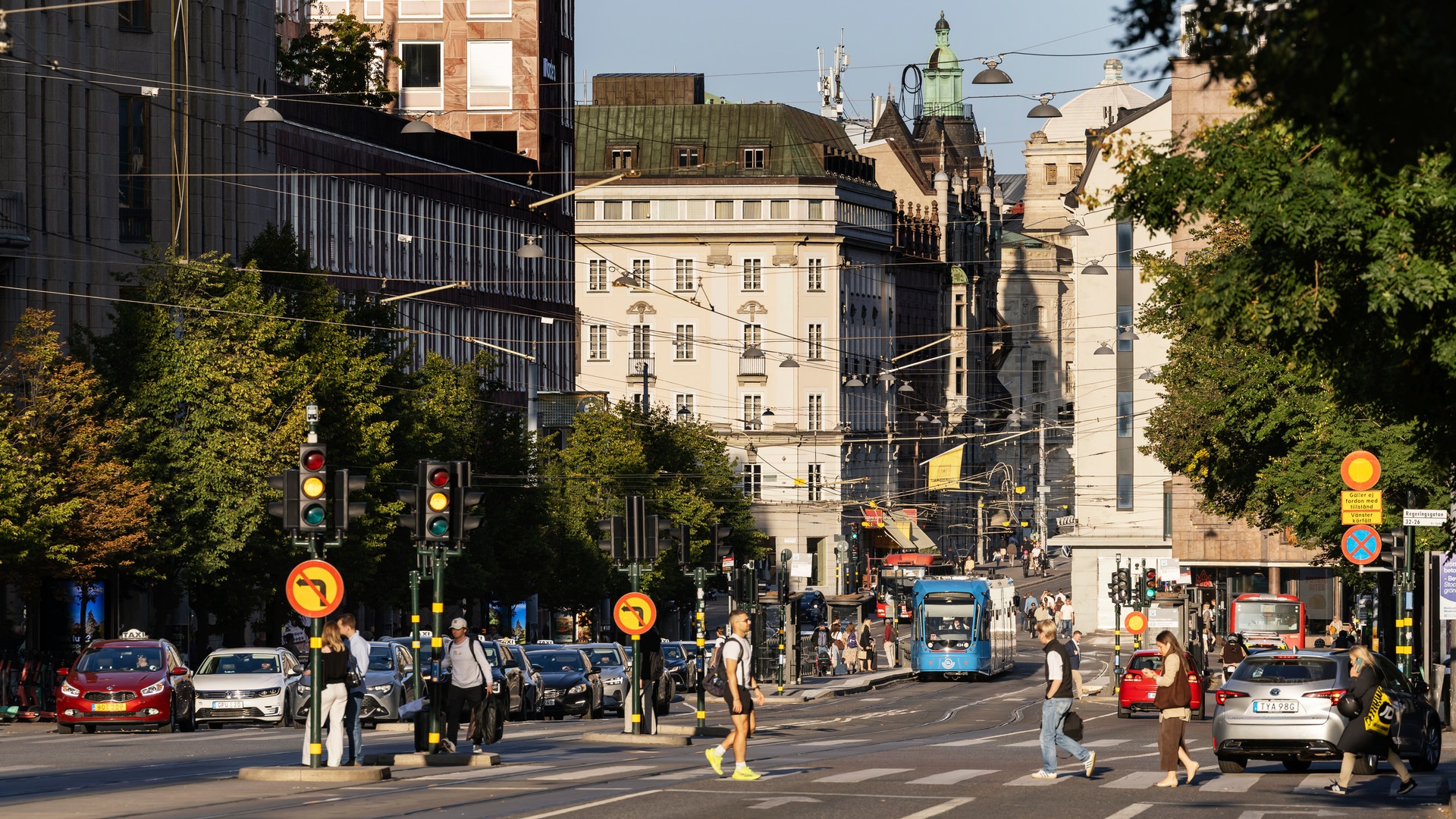 En stadsgata i en europeisk stad med klassiska byggnader, gågränd med fotgängare, trafikljus och gröna träd längs vägen.