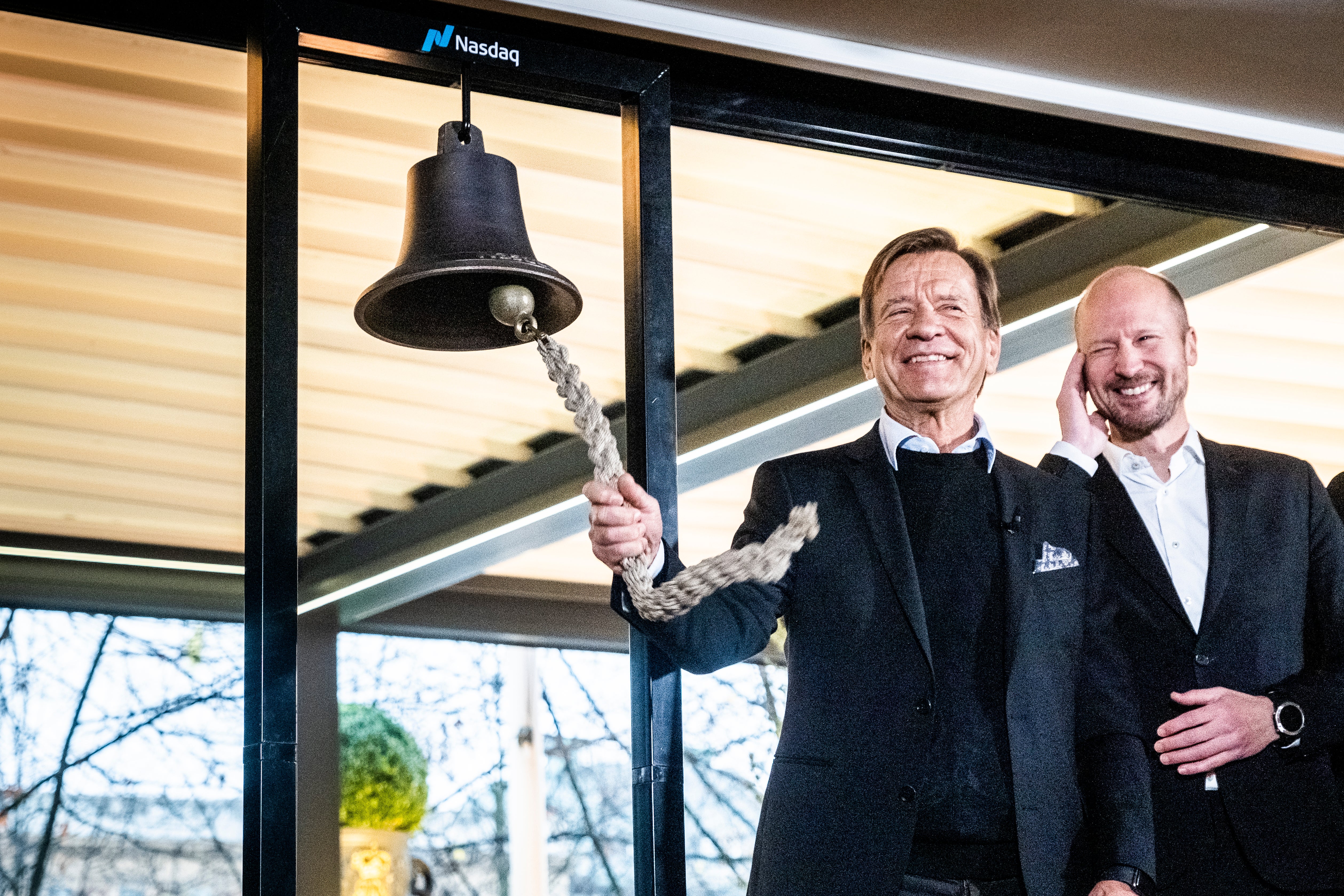 Två leende män i kostym vid en stor ringklocka märkt Nasdaq. Mannen till vänster drar i klockans rep.