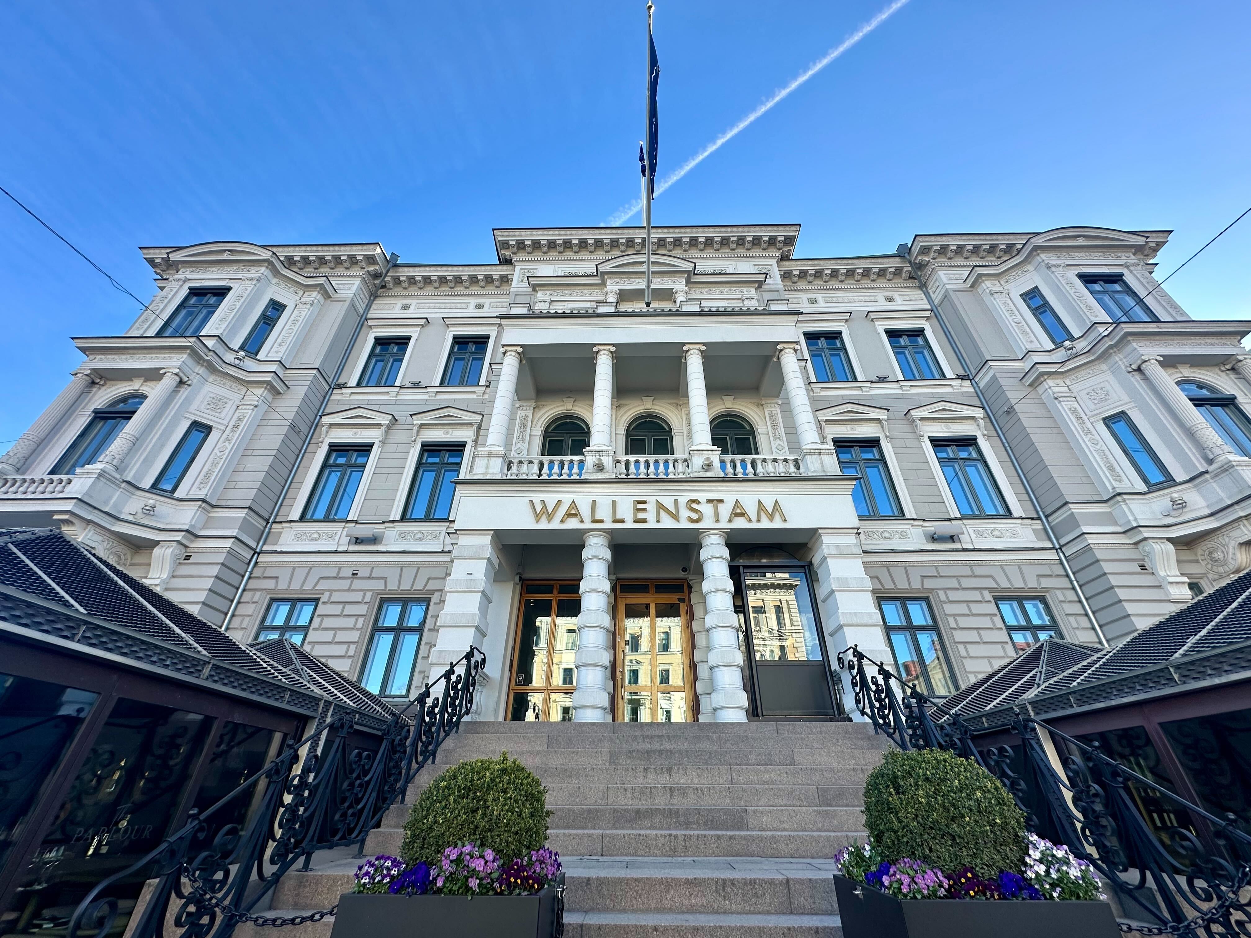 En klassisk vit stenhus med flera våningar och stora fönster. Byggnaden har kolonner vid huvudentrén och texten Wallenstam på fasaden. Framför huset står blomkrukor med blommor.