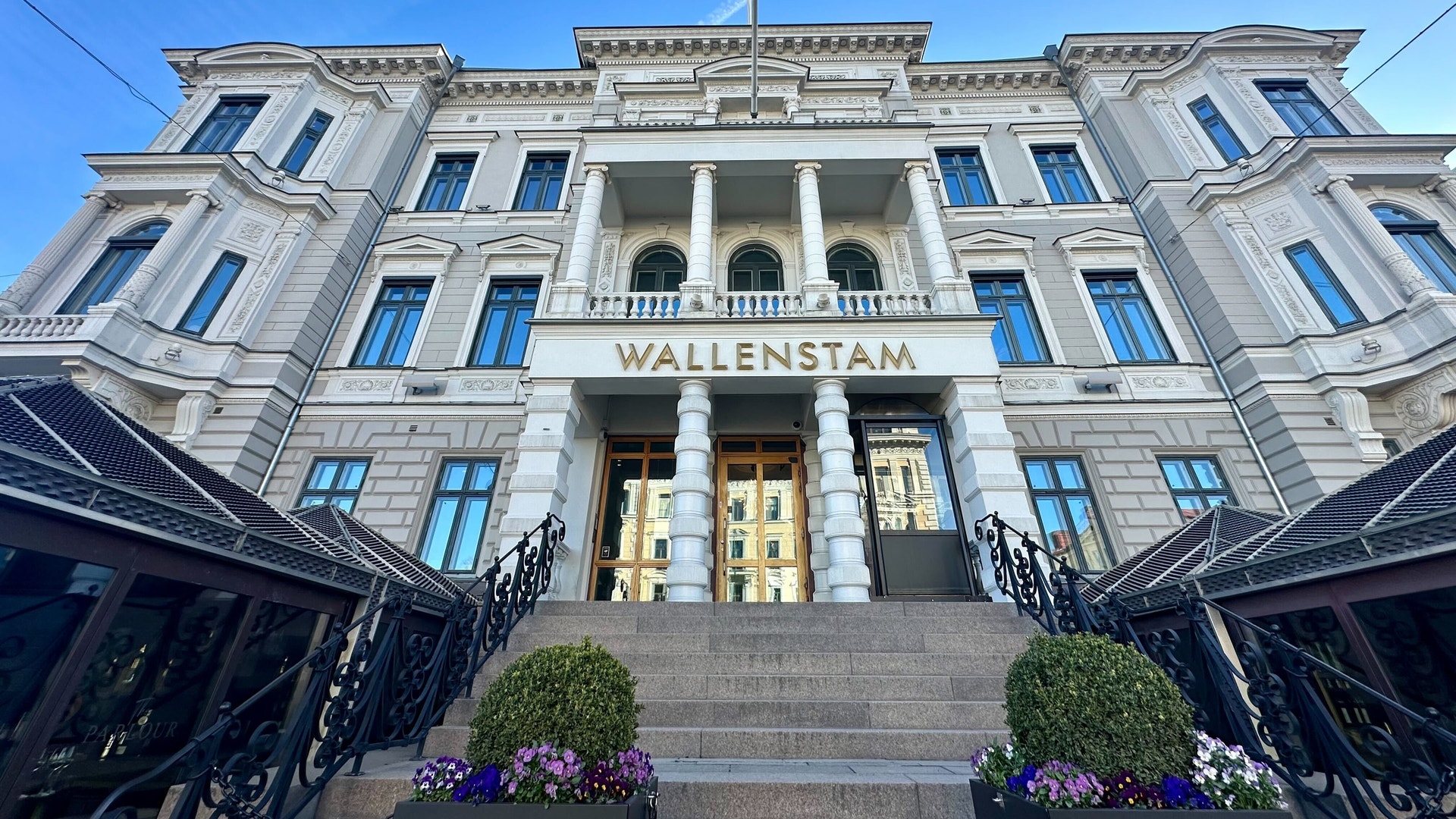 En klassisk vit stenhus med flera våningar och stora fönster. Byggnaden har kolonner vid huvudentrén och texten Wallenstam på fasaden. Framför huset står blomkrukor med blommor.