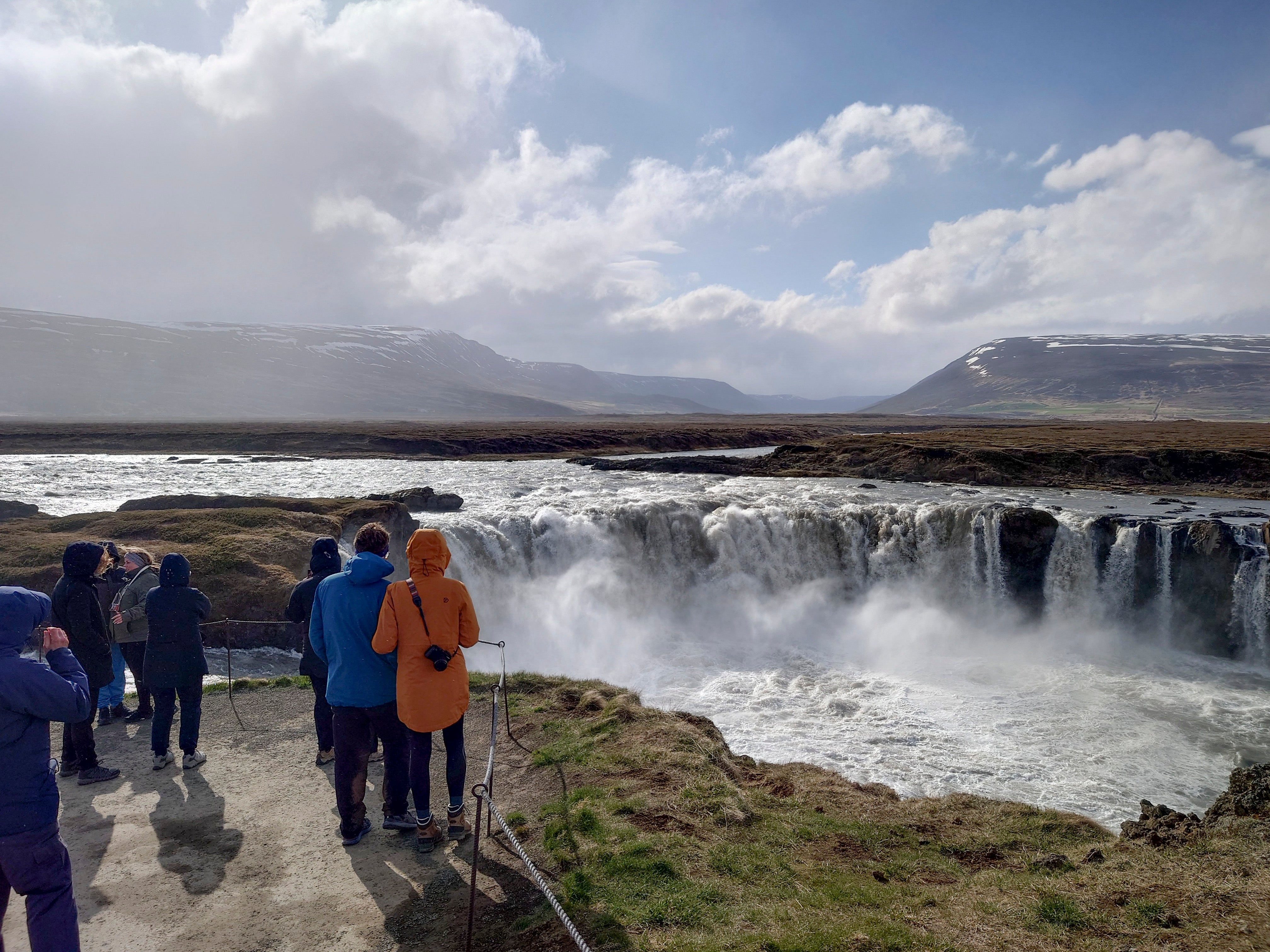 En kraftig vattenfall i Islands landskap. Turister i färgglada jackor står på en klippa och betraktar det brusande vattnet. Snöklädda berg syns i fjärran under en molnig himmel.