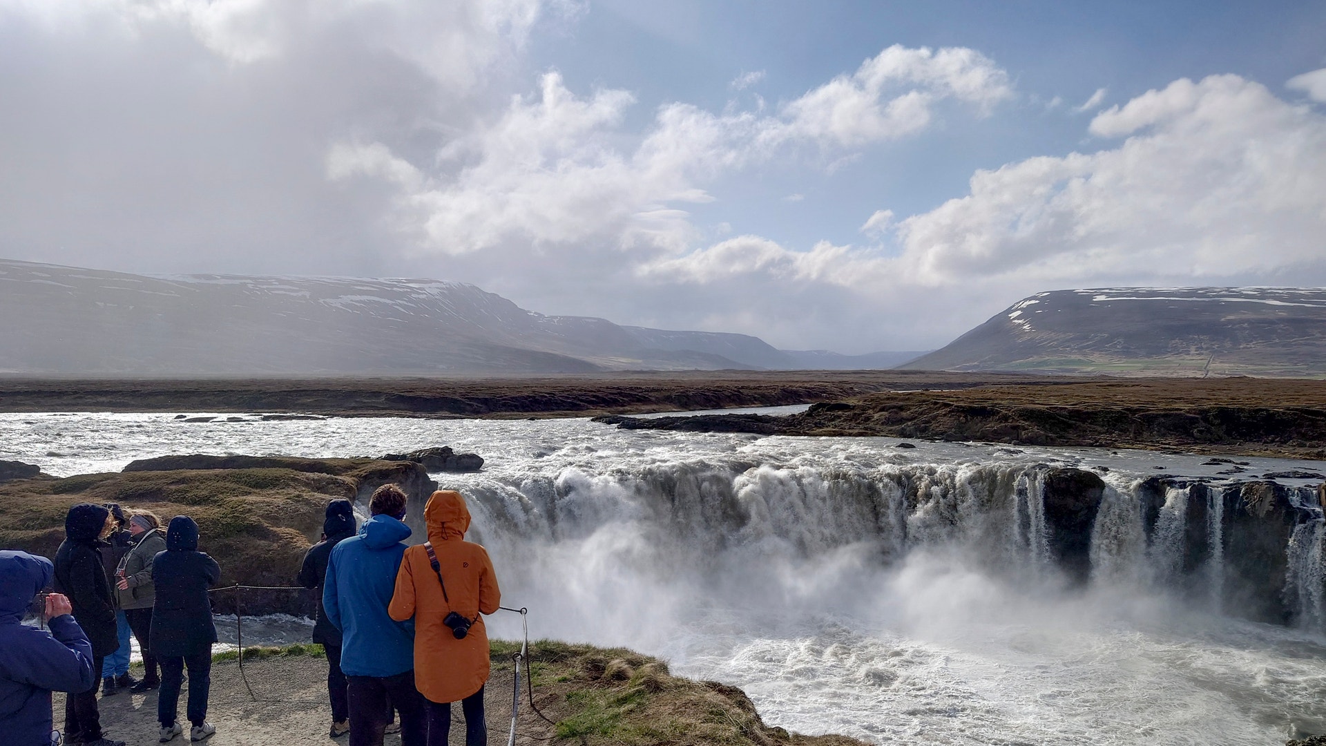 En kraftig vattenfall i Islands landskap. Turister i färgglada jackor står på en klippa och betraktar det brusande vattnet. Snöklädda berg syns i fjärran under en molnig himmel.