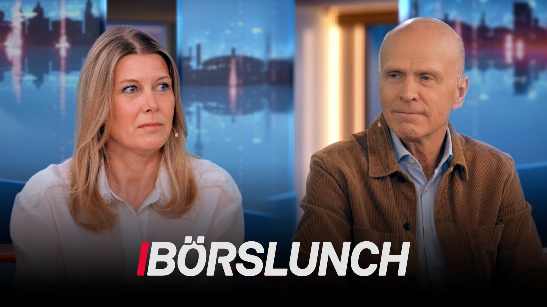Två personer sitter framför en stadshorisont i TV-programmet Börslunch. En kvinna i ljus skjorta och en man i brun jacka presenterar programmet tillsammans.