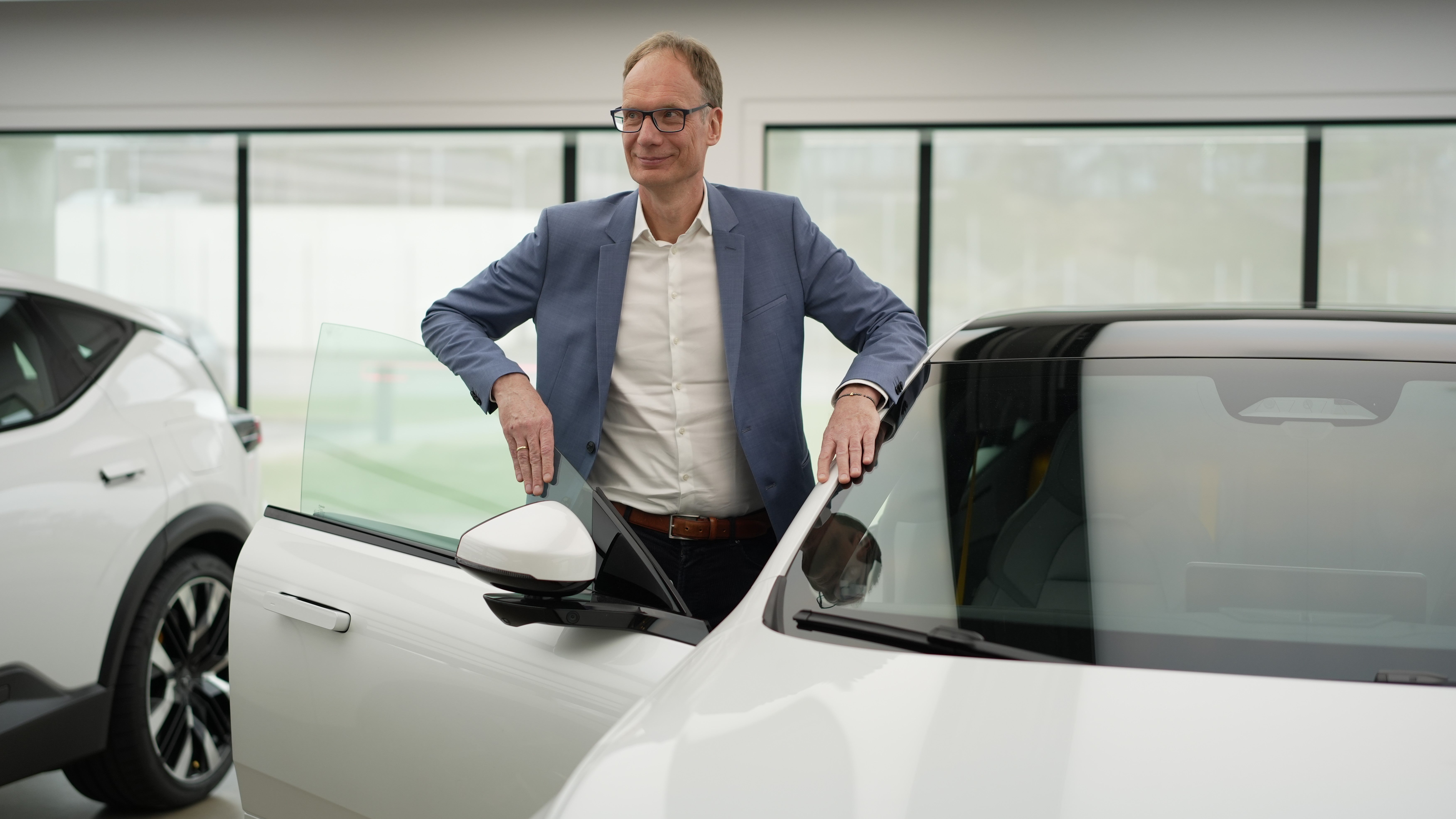 Man i blå kavaj står bredvid en vit bil och poserar för kameran.