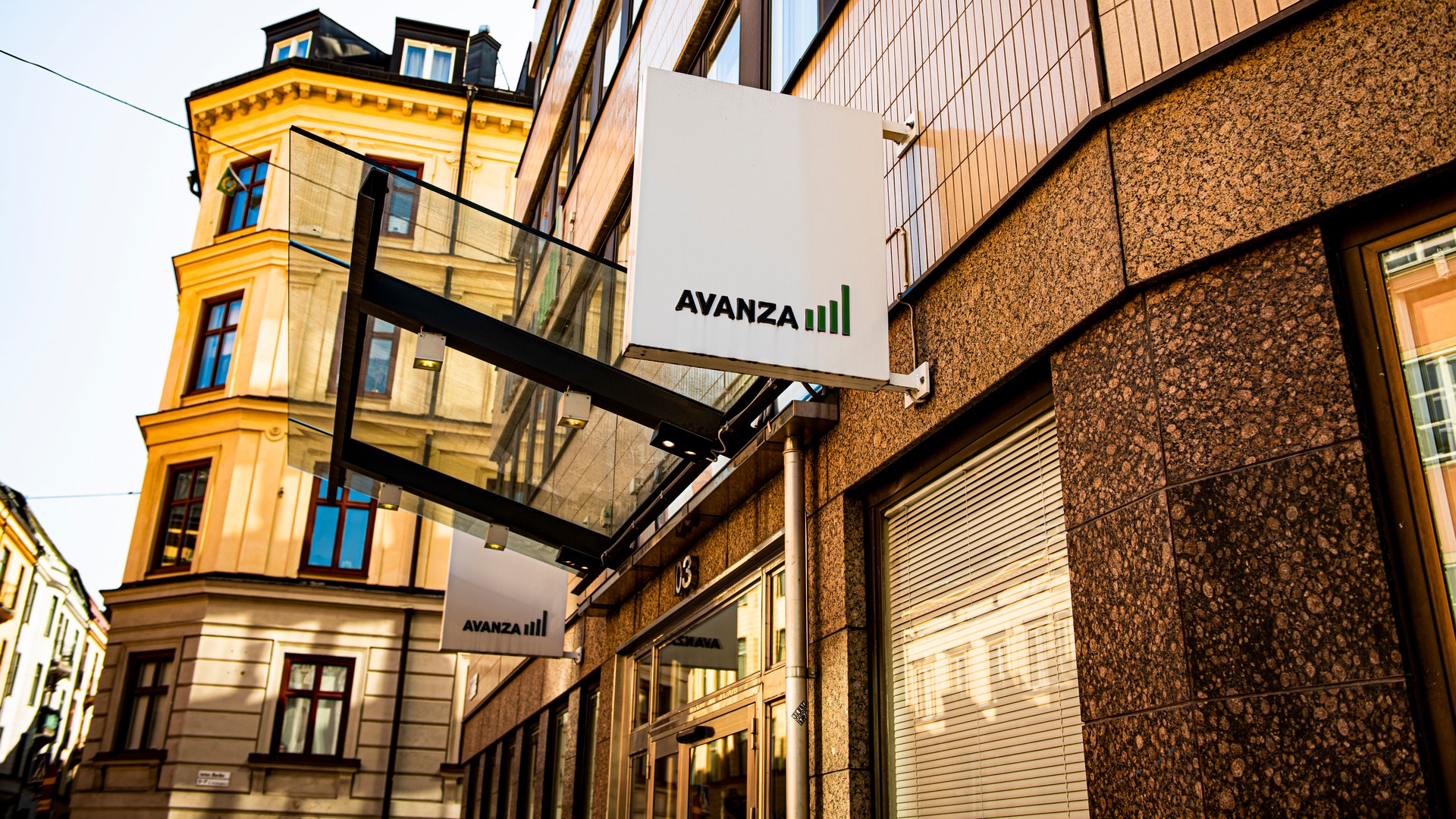 Gatuscen med en gul klassisk byggnad och moderna byggnader. En svart metallkonstruktion sträcker sig över gatan med en vit skylt märkt AVANZA.