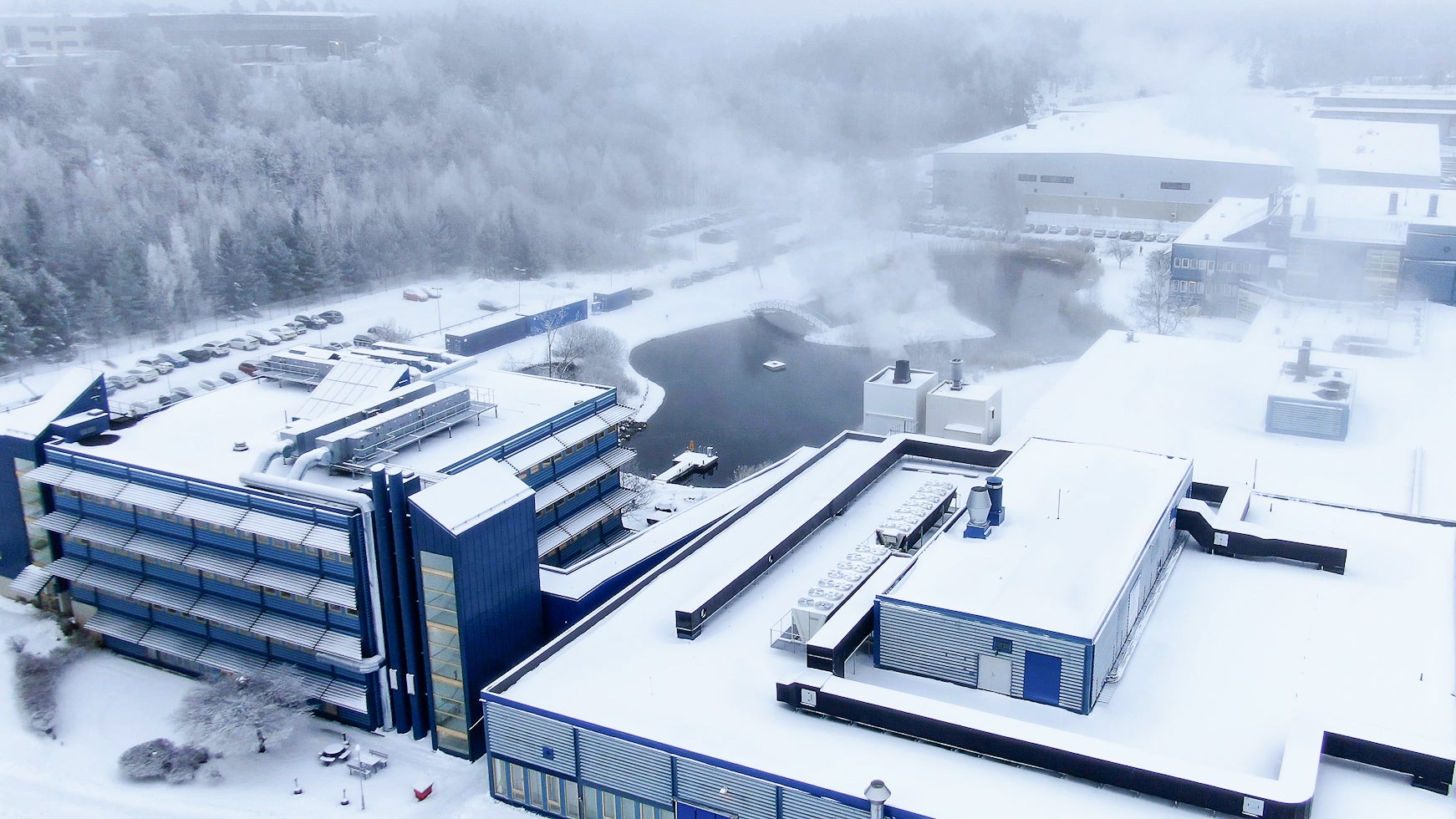 Luftfoto av en industrianläggning med blå och vita byggnader omgiven av snö och barrskog.