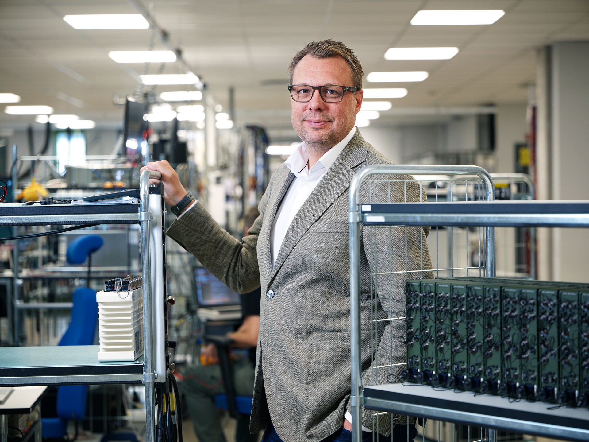 En man i kavaj står i en fabrikslokal bland vagnar med elektronisk utrustning.