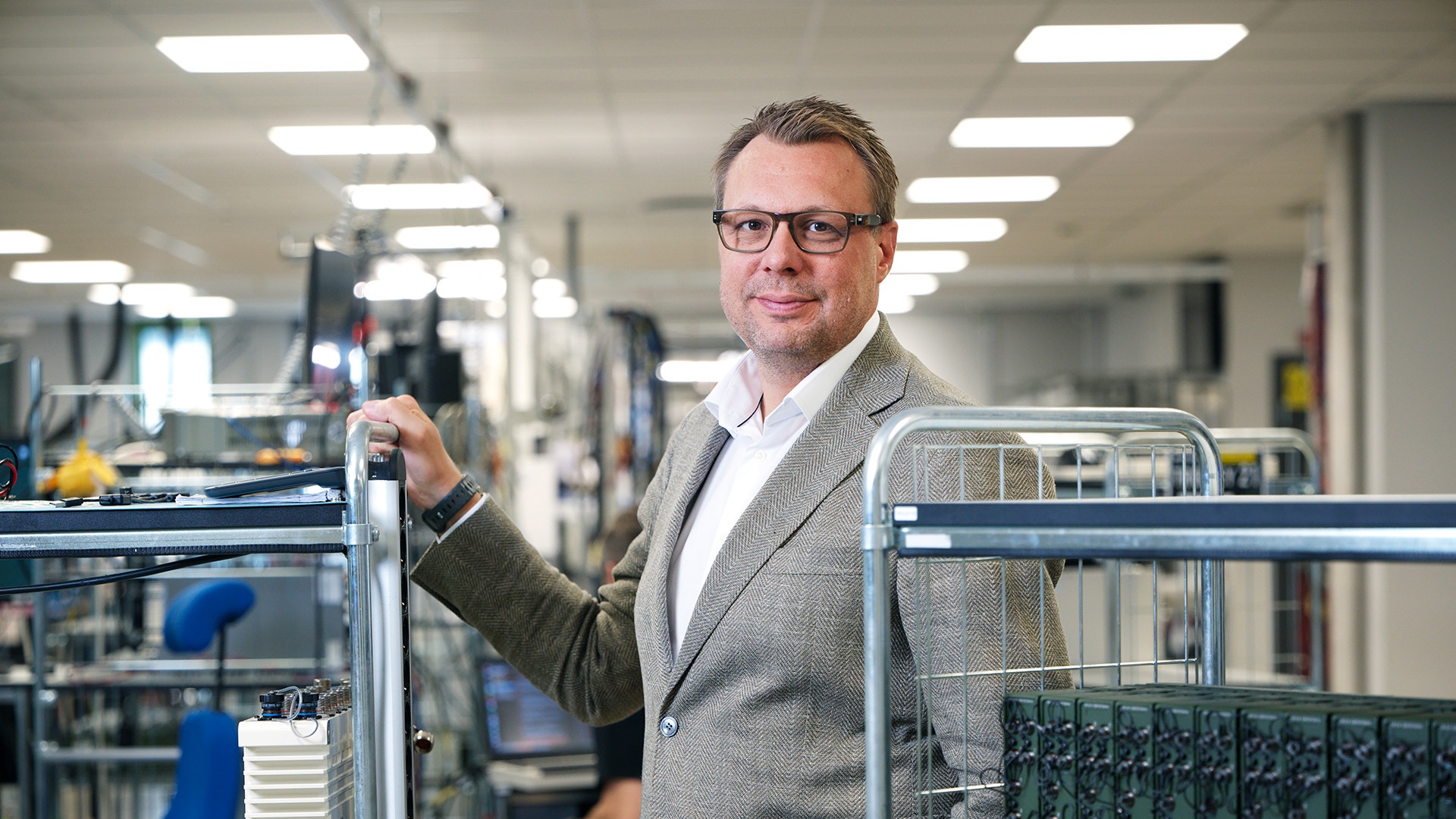 En man i kavaj står i en fabrikslokal bland vagnar med elektronisk utrustning.