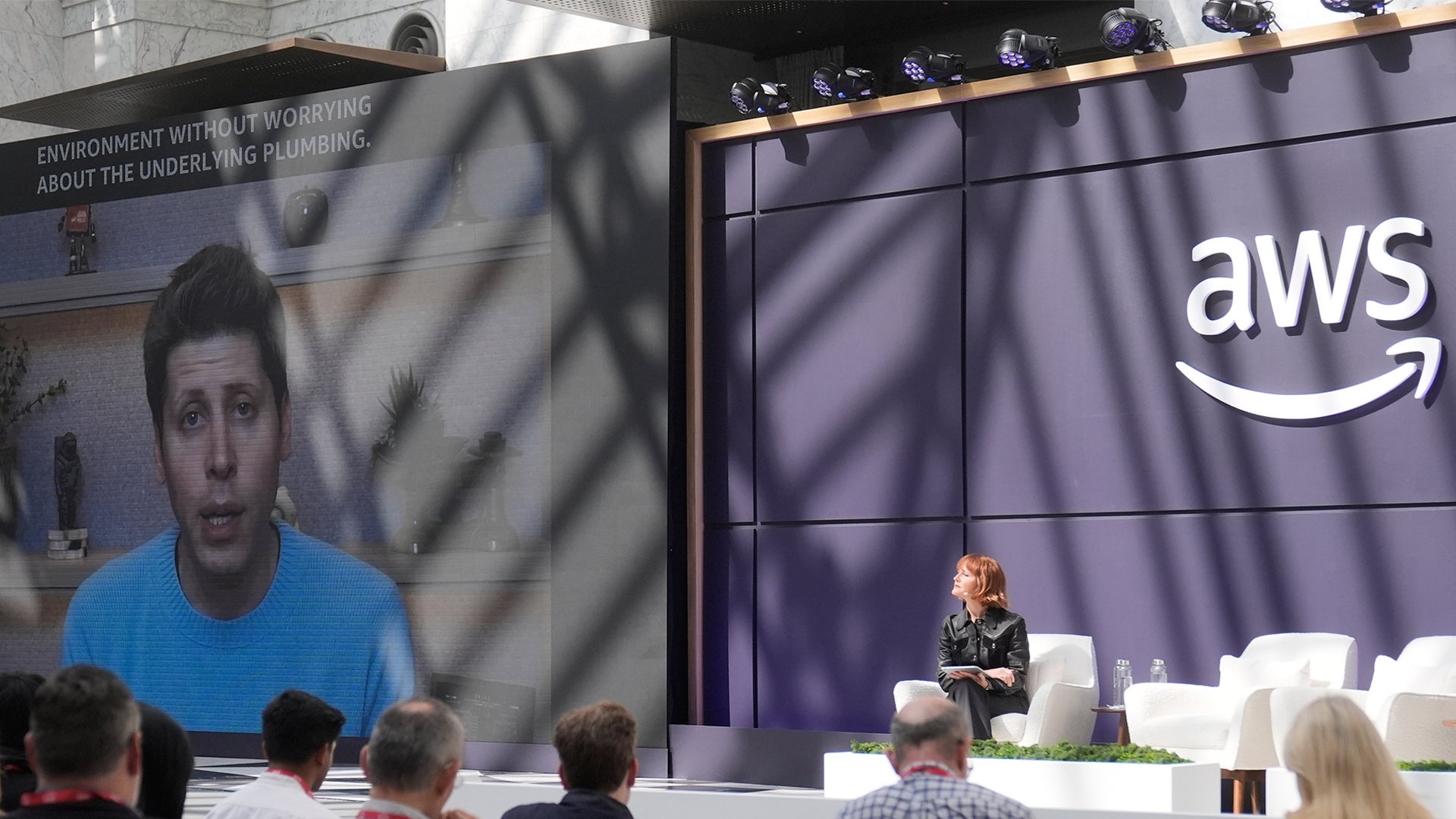 En person presenterar på en konferens framför en stor skärm med AWS-logotyp. Publiken sitter och lyssnar på presentationen.