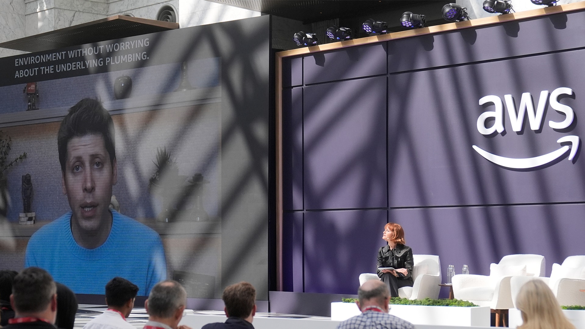 En person presenterar på en konferens framför en stor skärm med AWS-logotyp. Publiken sitter och lyssnar på presentationen.