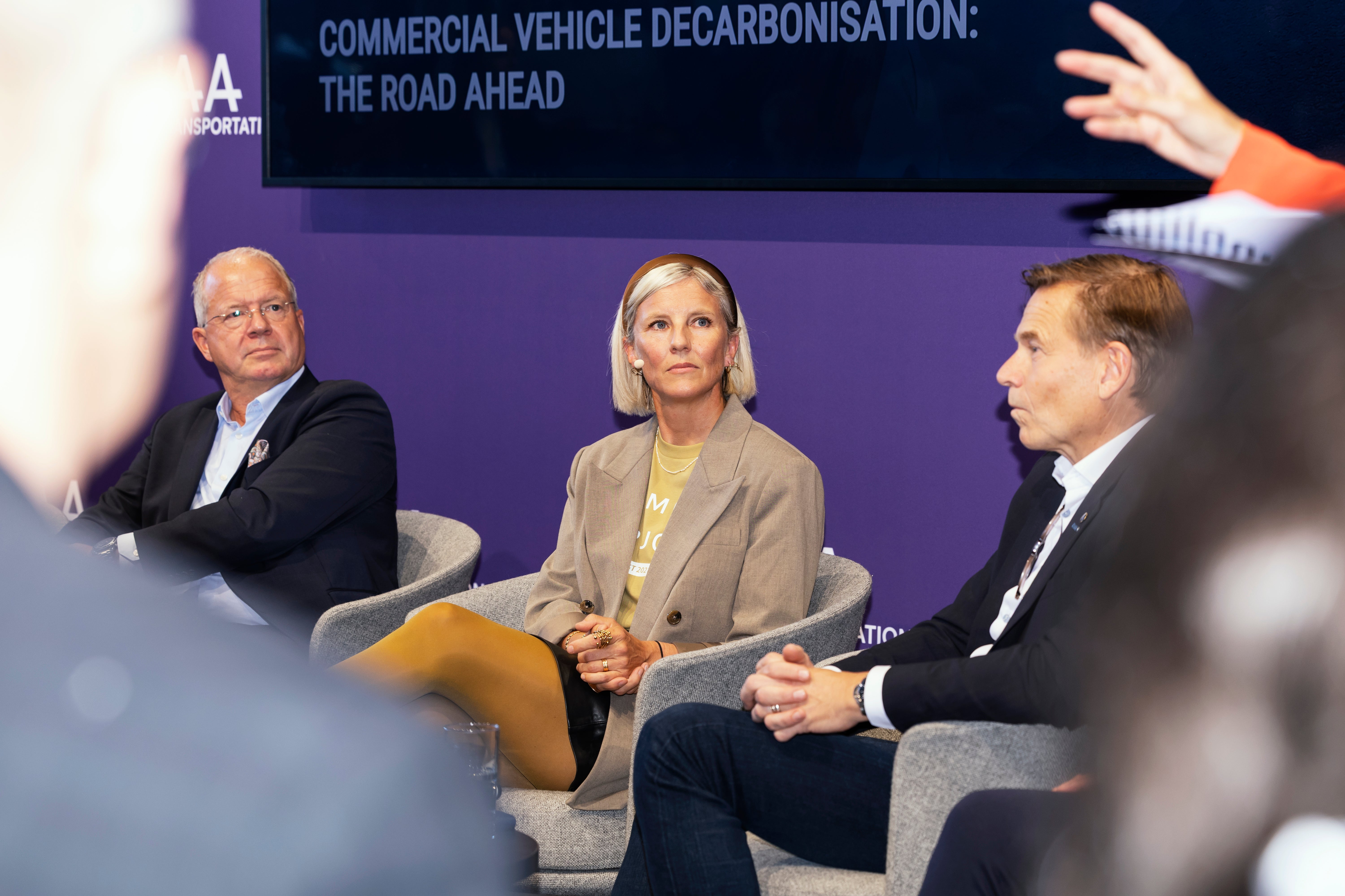 Tre personer sitter i stolar på scen under en paneldiskussion, med en skärm bakom dem som visar rubriken "Commercial Vehicle Decarbonisation: The Road Ahead".