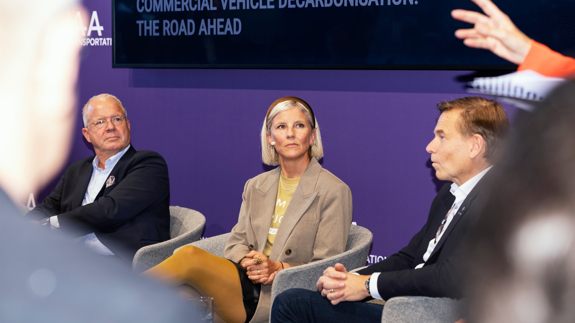 Tre personer sitter i stolar på scen under en paneldiskussion, med en skärm bakom dem som visar rubriken "Commercial Vehicle Decarbonisation: The Road Ahead".