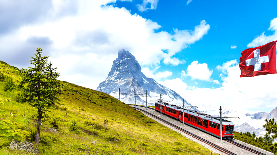 En genrebild på Schweiz uppe i bergen, där ett rött tåg åker förbi landets flagga.