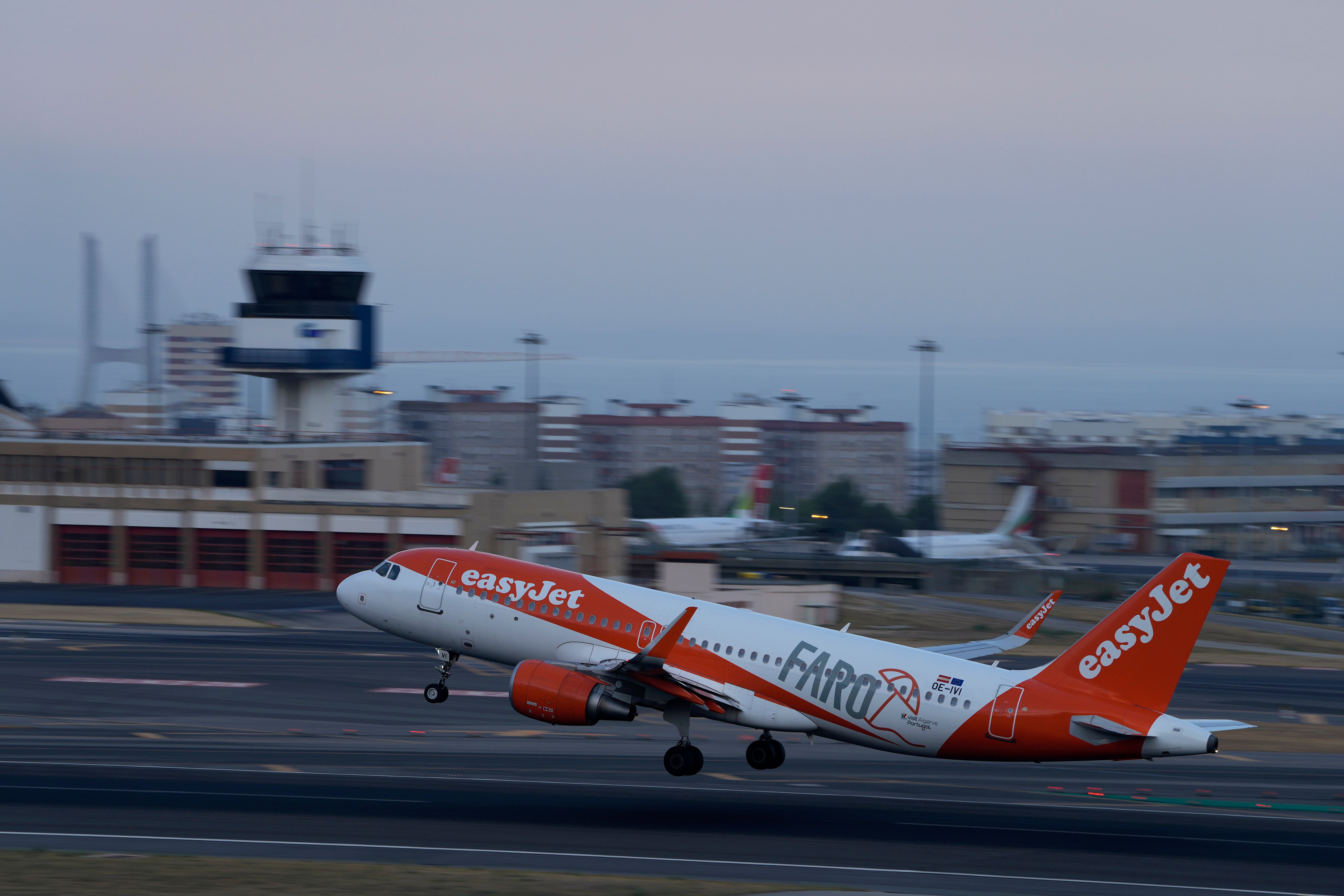 Ett easyJet-flygplan med orange och vit färg lyfter från en flygplats. Flygplatsterminalen och kontrolltornet syns i bakgrunden.