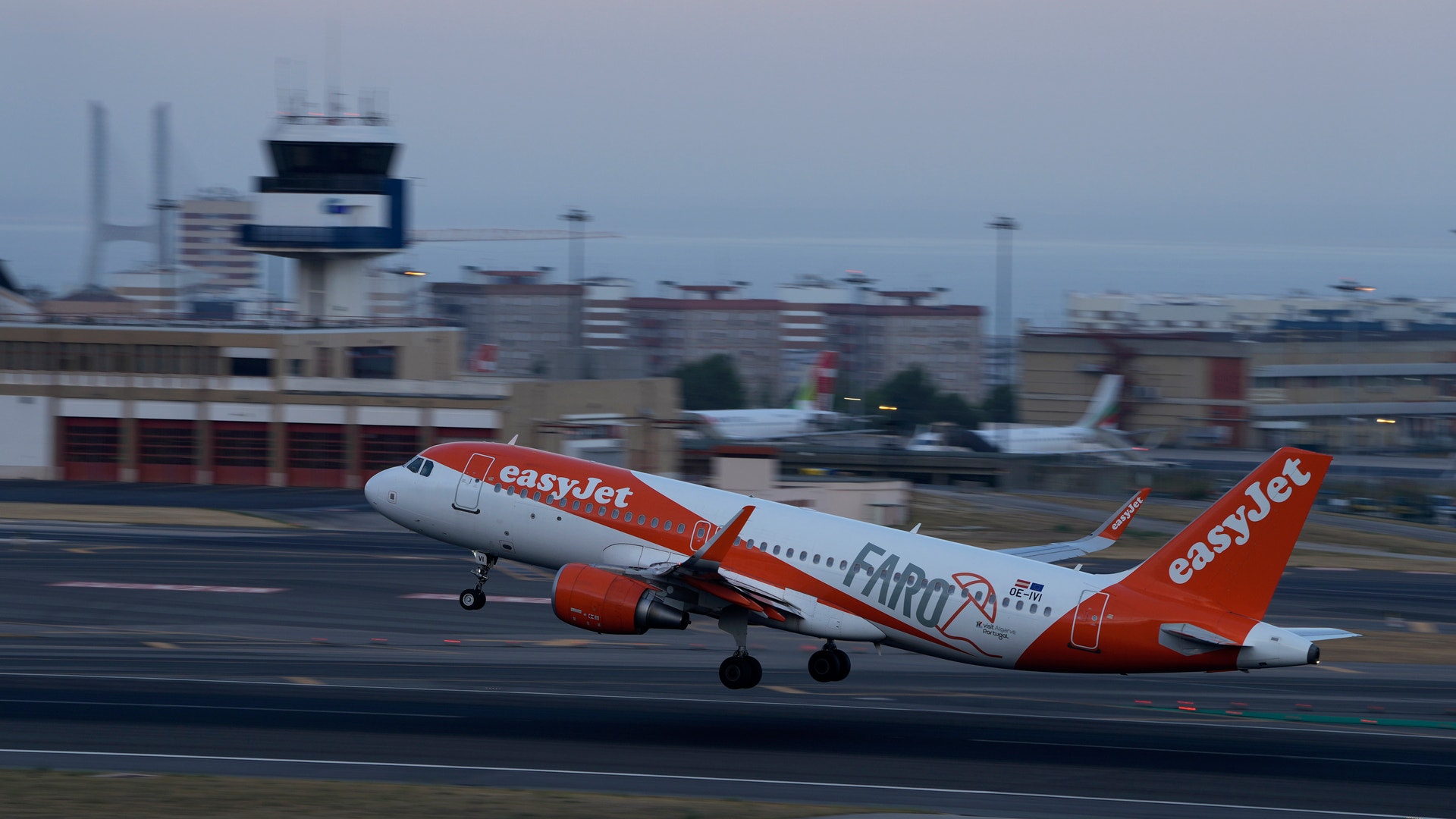 Ett easyJet-flygplan med orange och vit färg lyfter från en flygplats. Flygplatsterminalen och kontrolltornet syns i bakgrunden.