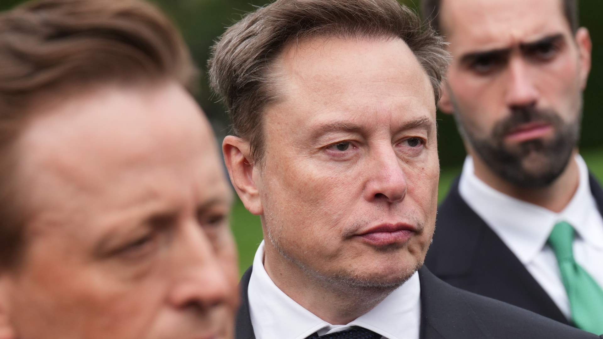 Närbild på tre allvarliga män i kostym utomhus. Mannen i mitten tittar åt sidan.