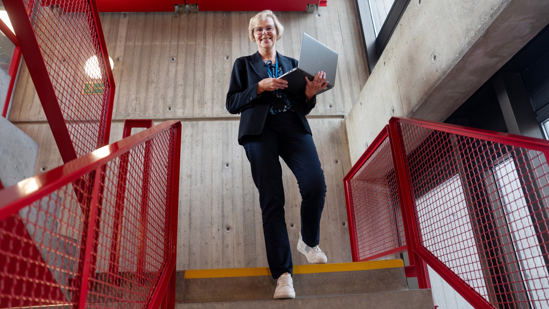 Leende kvinna i mörk kavaj och byxor går ner för en trappa med röda räcken, med en laptop i handen.