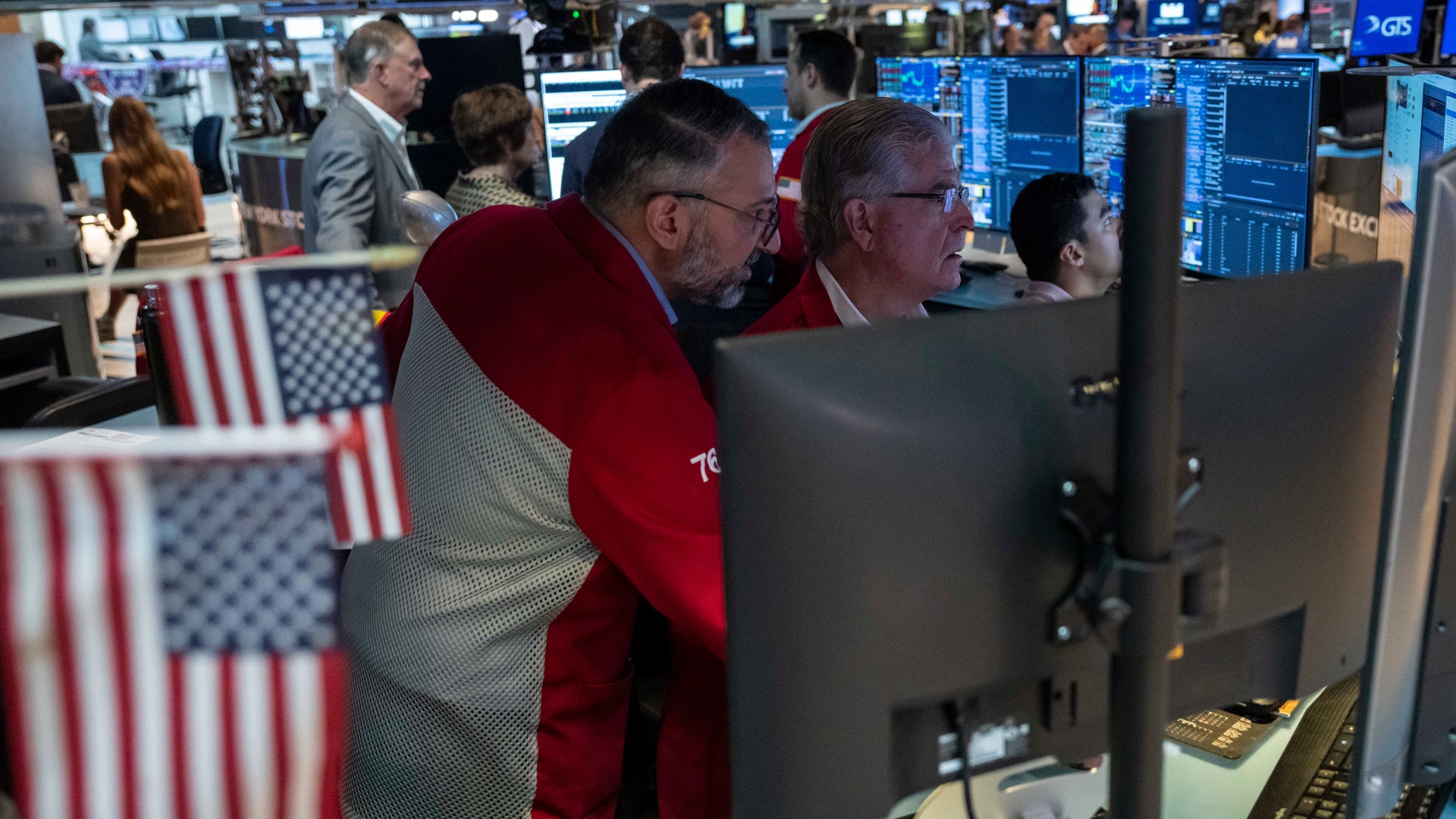 Handlare arbetar vid dataskärmar på New York Stock Exchange. Amerikanska flaggor syns i förgrunden och börsgolvet är fullt av människor och handelsstationer.