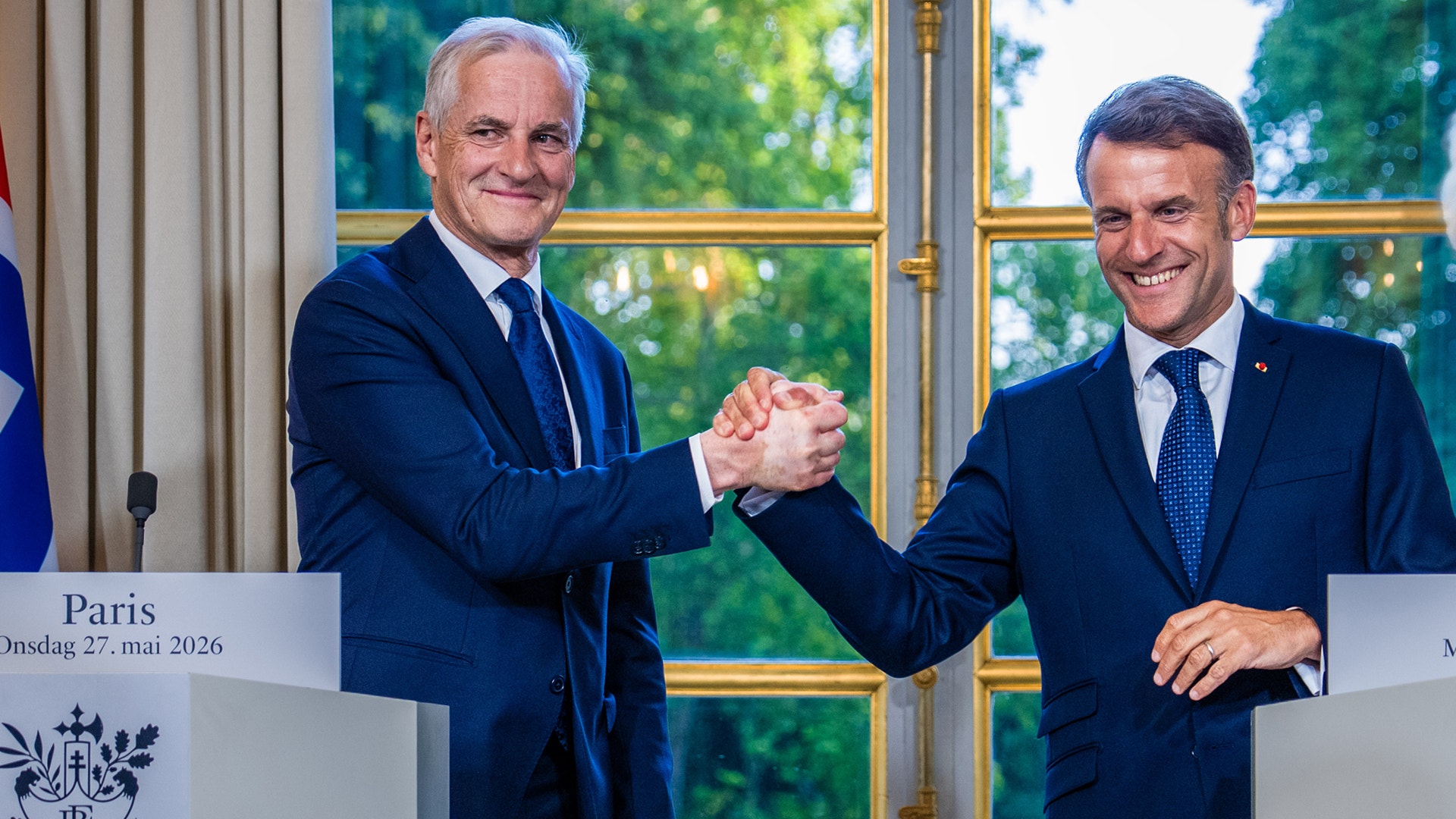 Två män i mörka kostymer skakar hand vid ett officiellt möte i Paris. De står framför ett fönster med gyllene ramar.