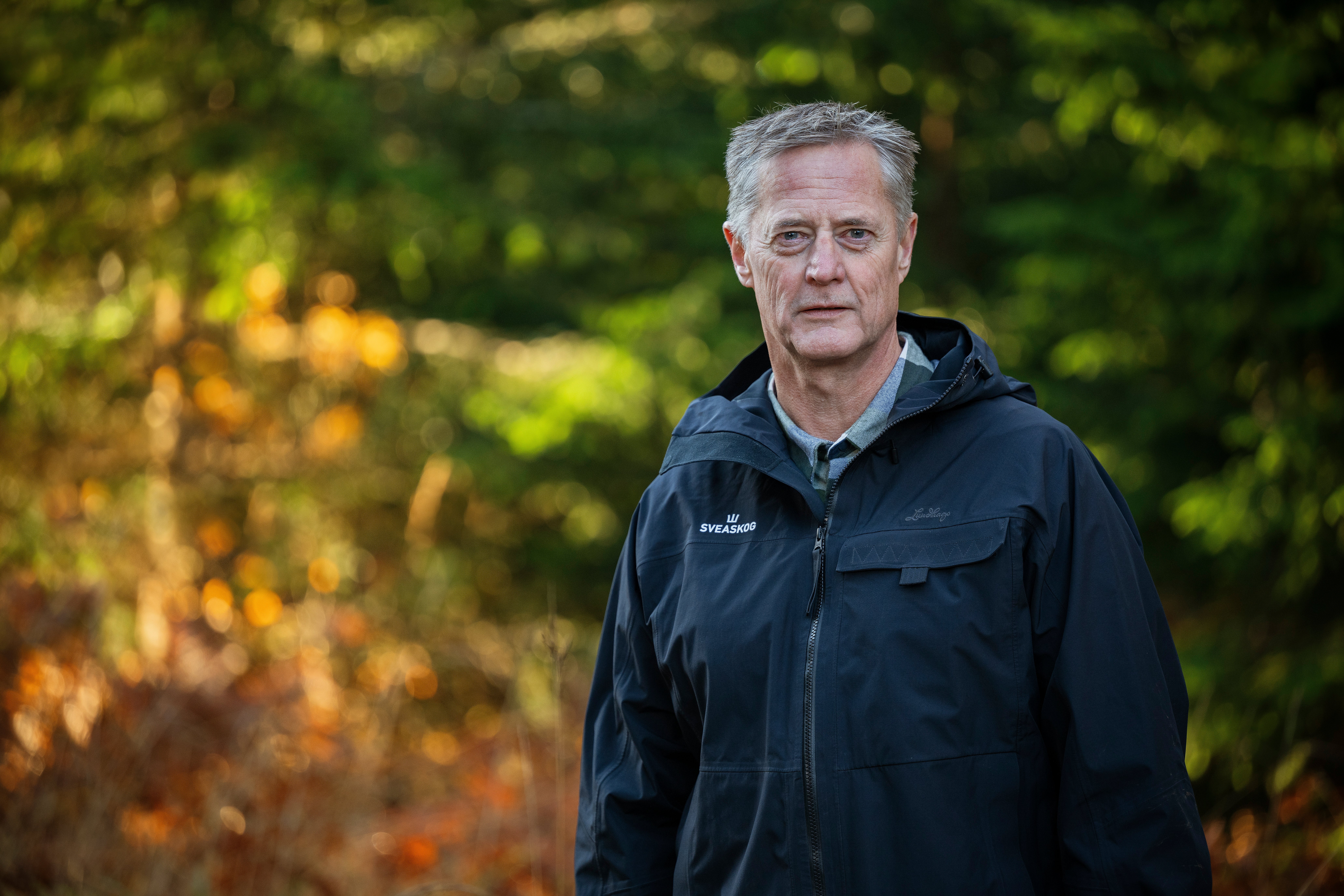 Porträtt av en äldre man med grått hår i mörk jacka som står utomhus framför skog i höstfärger.